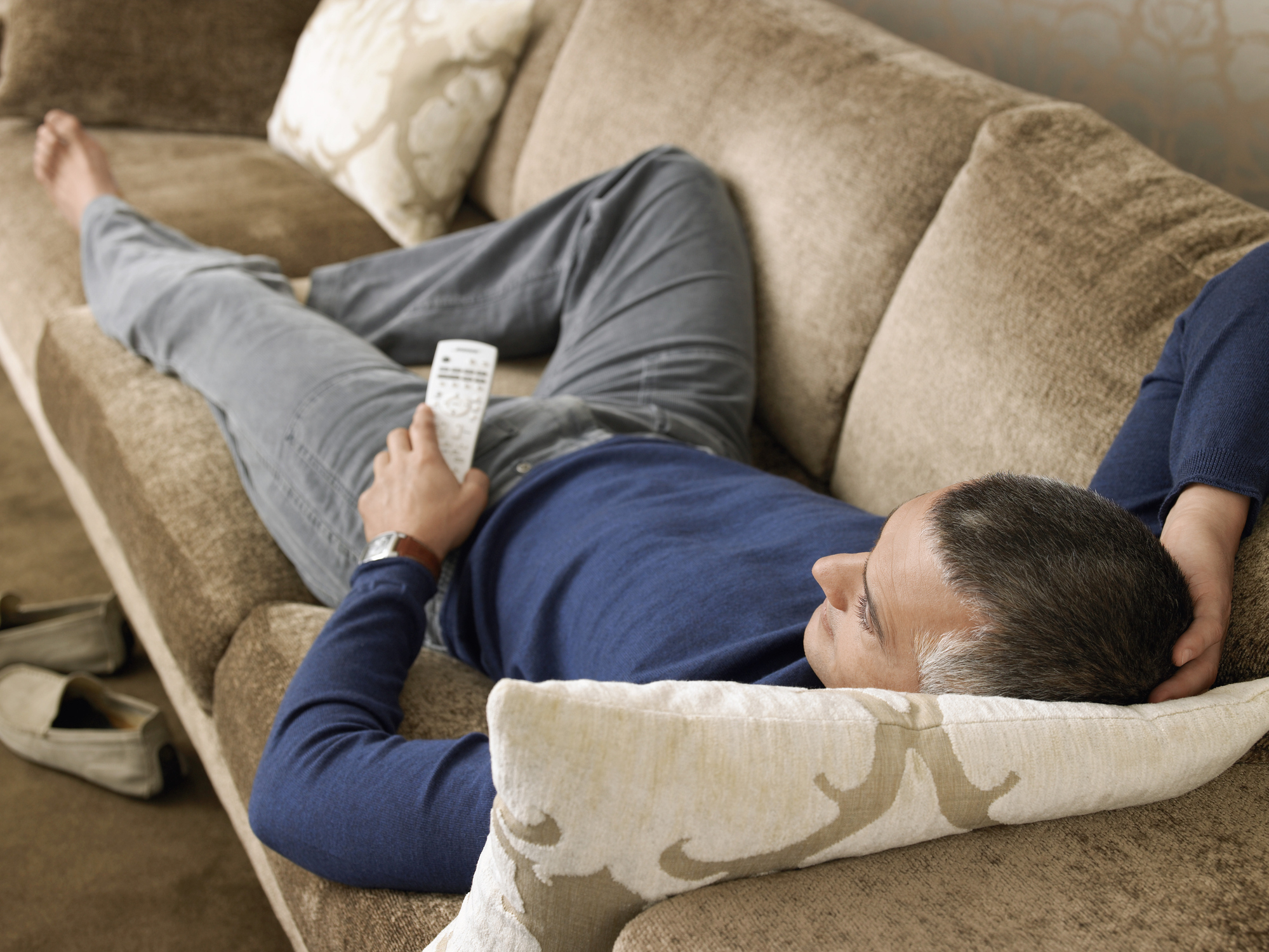 A person is lying on a couch, holding a remote control, and appears to be relaxed, possibly watching TV or resting. Shoes are on the floor nearby