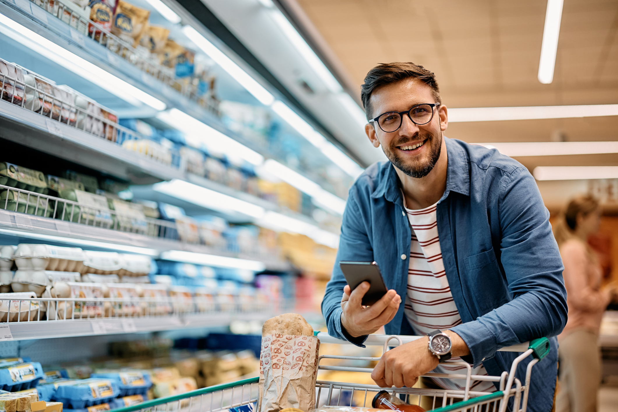 Man in a grocery store holding a smartphone, smiling next to a cart filled with groceries. Other shoppers in background