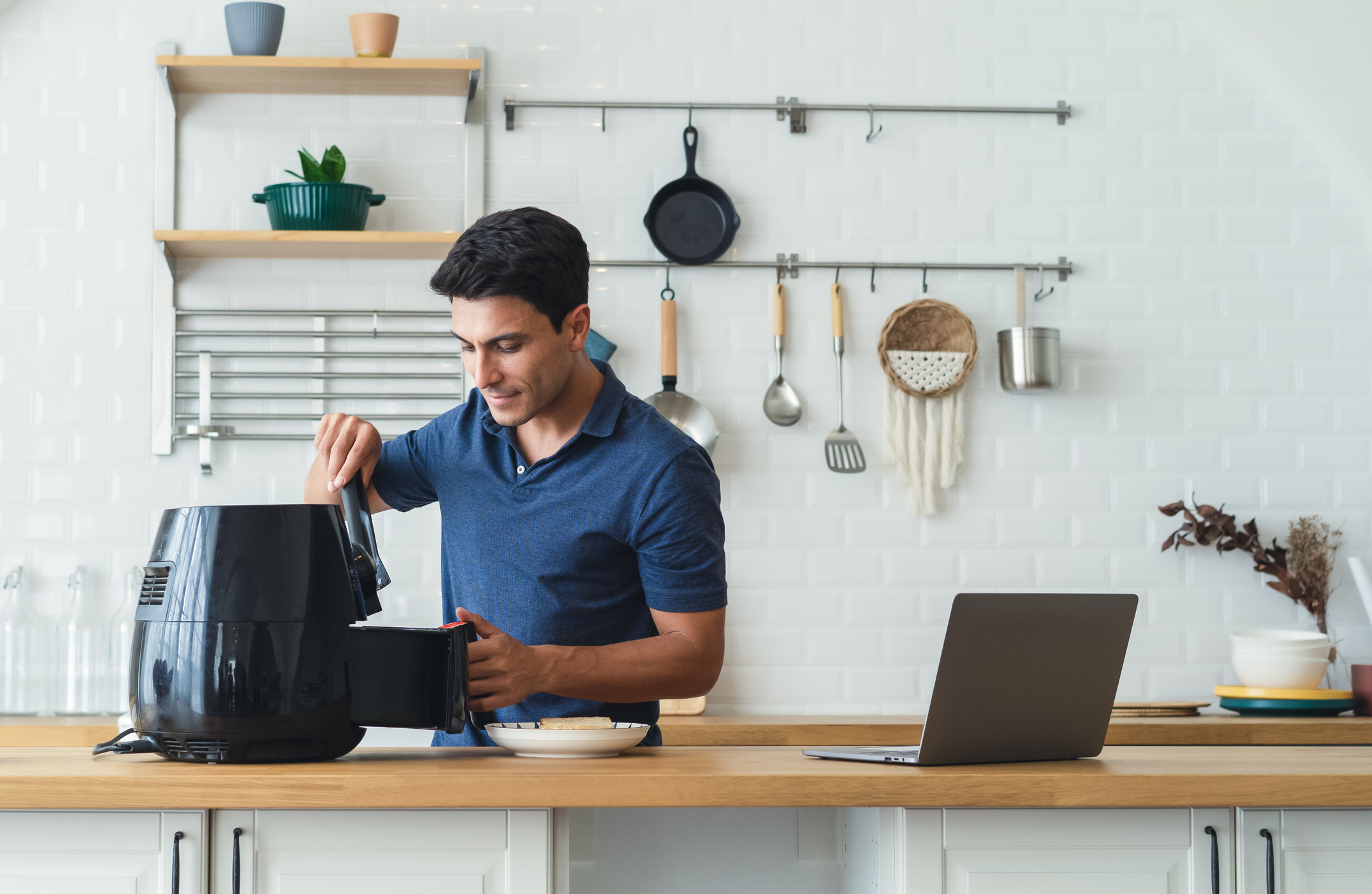 A man in a kitchen uses an air fryer while looking focused, with a laptop open on the counter beside him. Various kitchen utensils hang on the wall