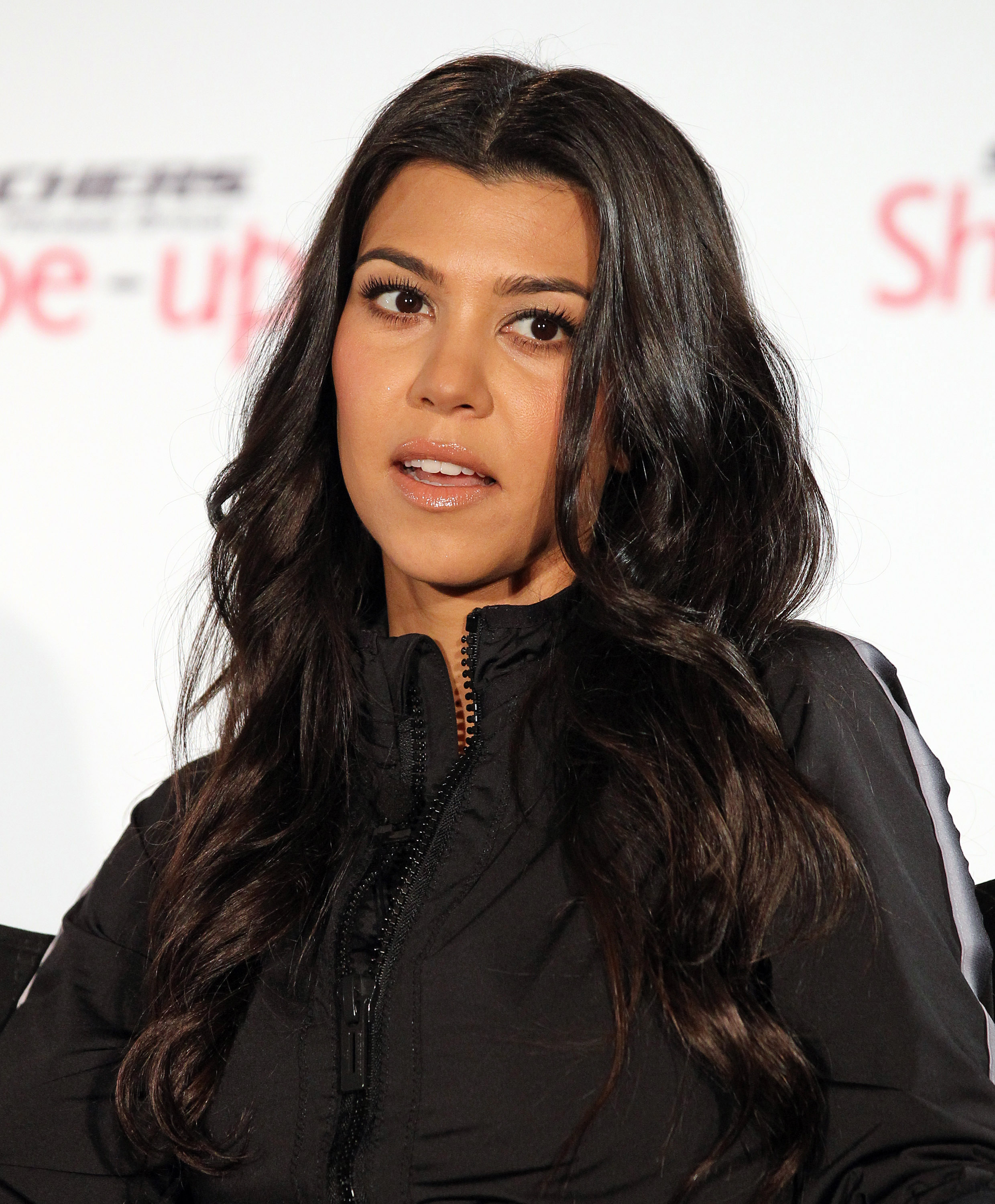 Kourtney Kardashian speaking at an event, wearing a casual jacket with wavy hair, looking slightly to the side