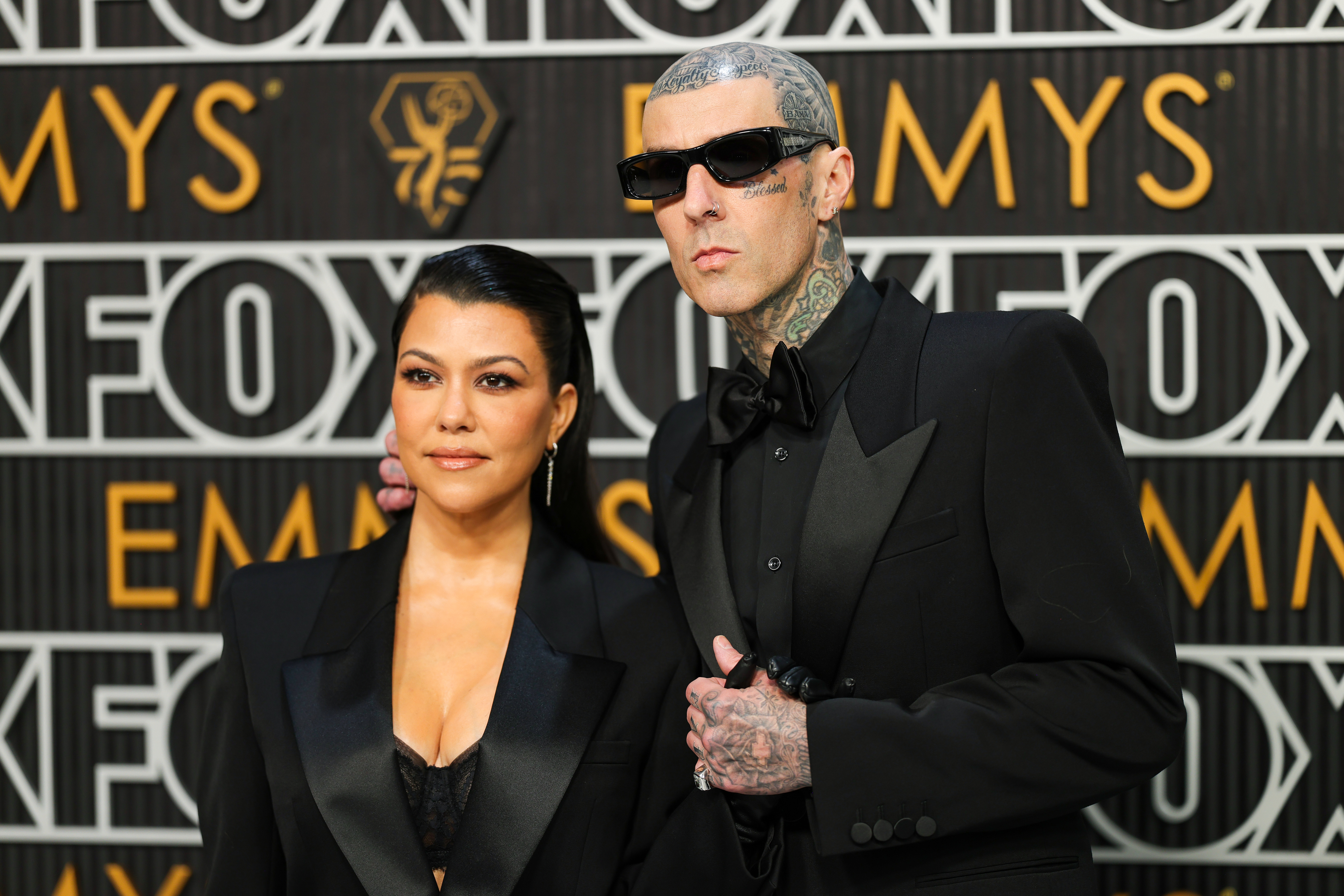 Kourtney Kardashian and Travis Barker on the red carpet at the Emmys. Kourtney is in a sleek outfit with a blazer, and Travis is in a dark suit with sunglasses