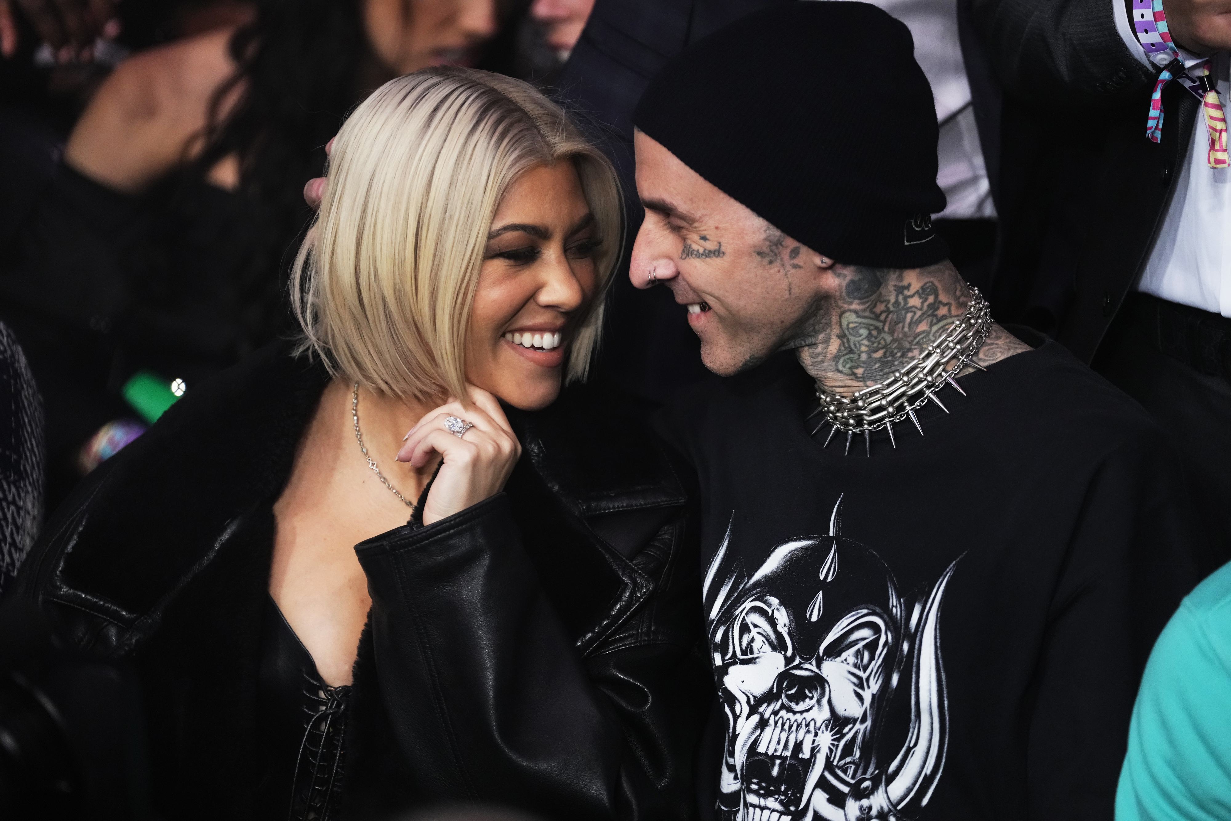 Kourtney Kardashian and Travis Barker smiling at each other. Kourtney has a sleek bob haircut and is in a plunging dark outfit. Travis wears a beanie and a spiked necklace