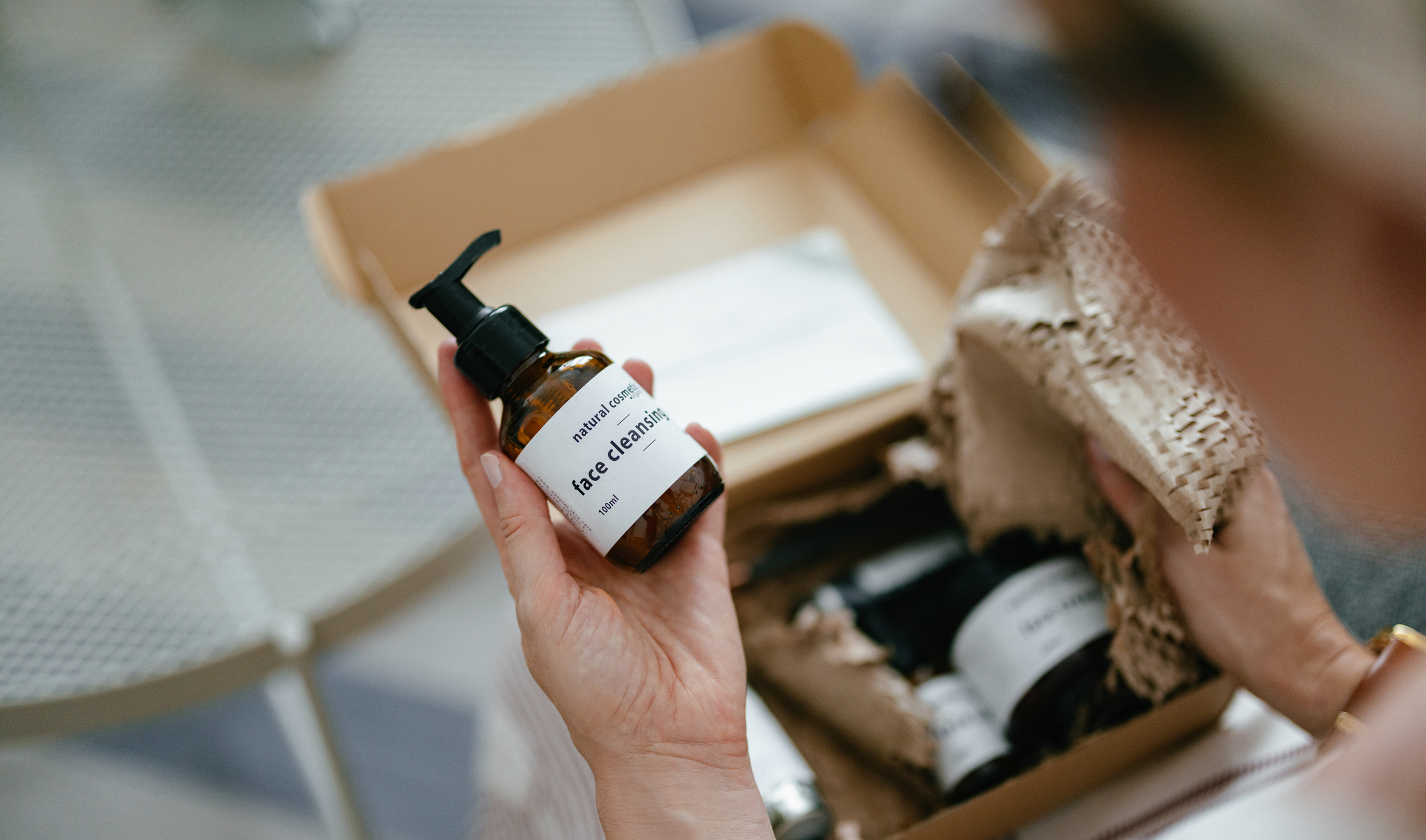 A hand holding a bottle labeled &quot;face cleanser&quot; next to an open cardboard box with packing material and other cosmetic items