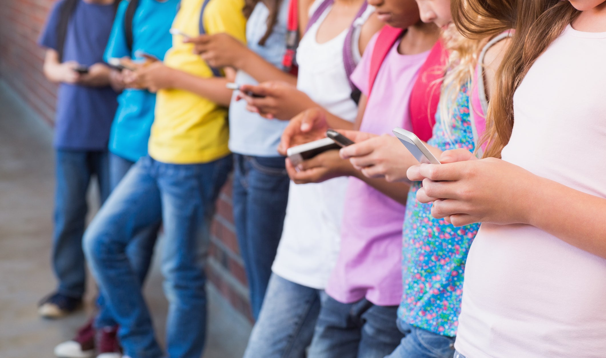 A group of kids leaning against a wall, all looking down at their smartphones, engrossed in their screens