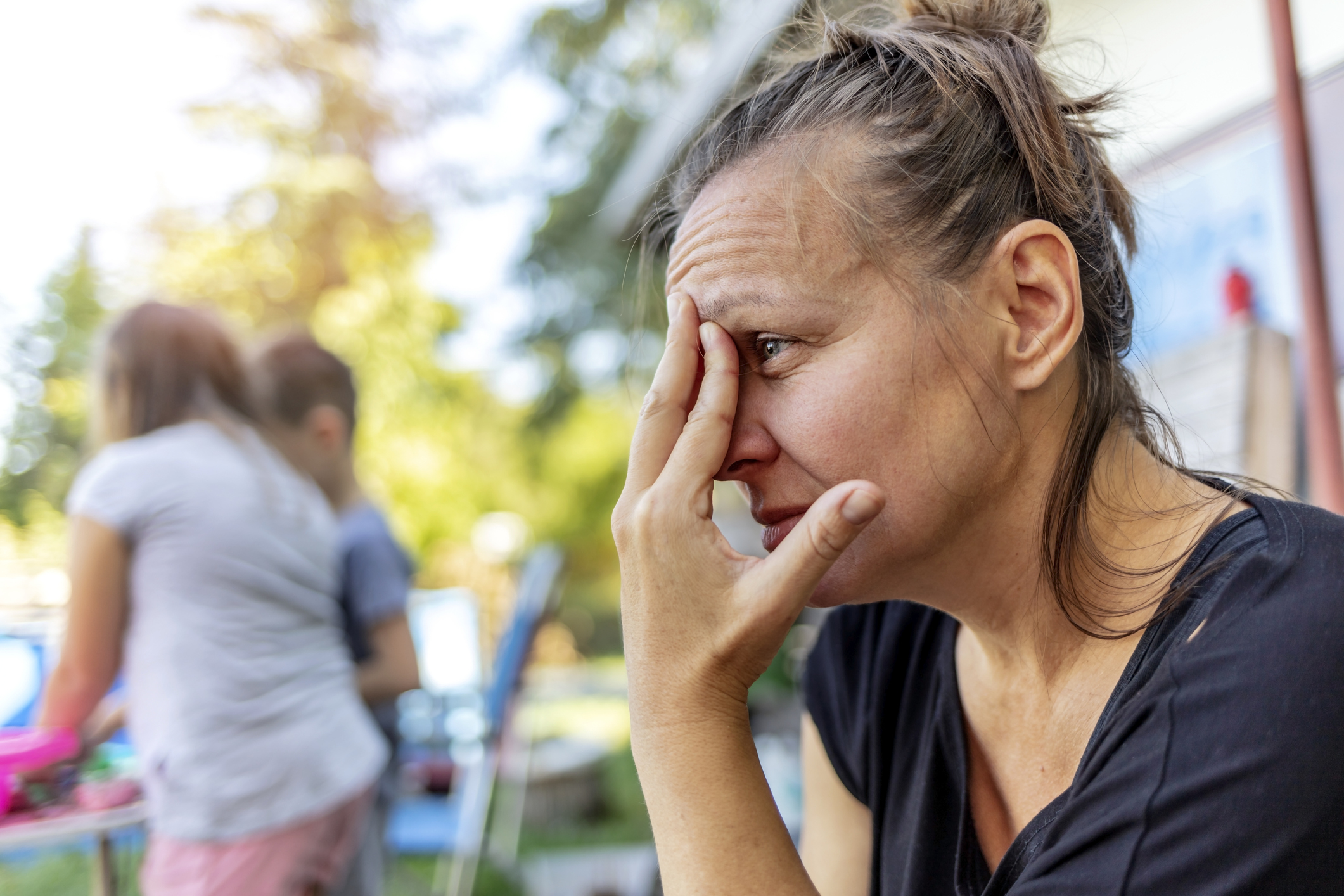 A woman looks stressed, covering part of her face with her hand. In the background, two children are playing in an outdoor setting. Names not available