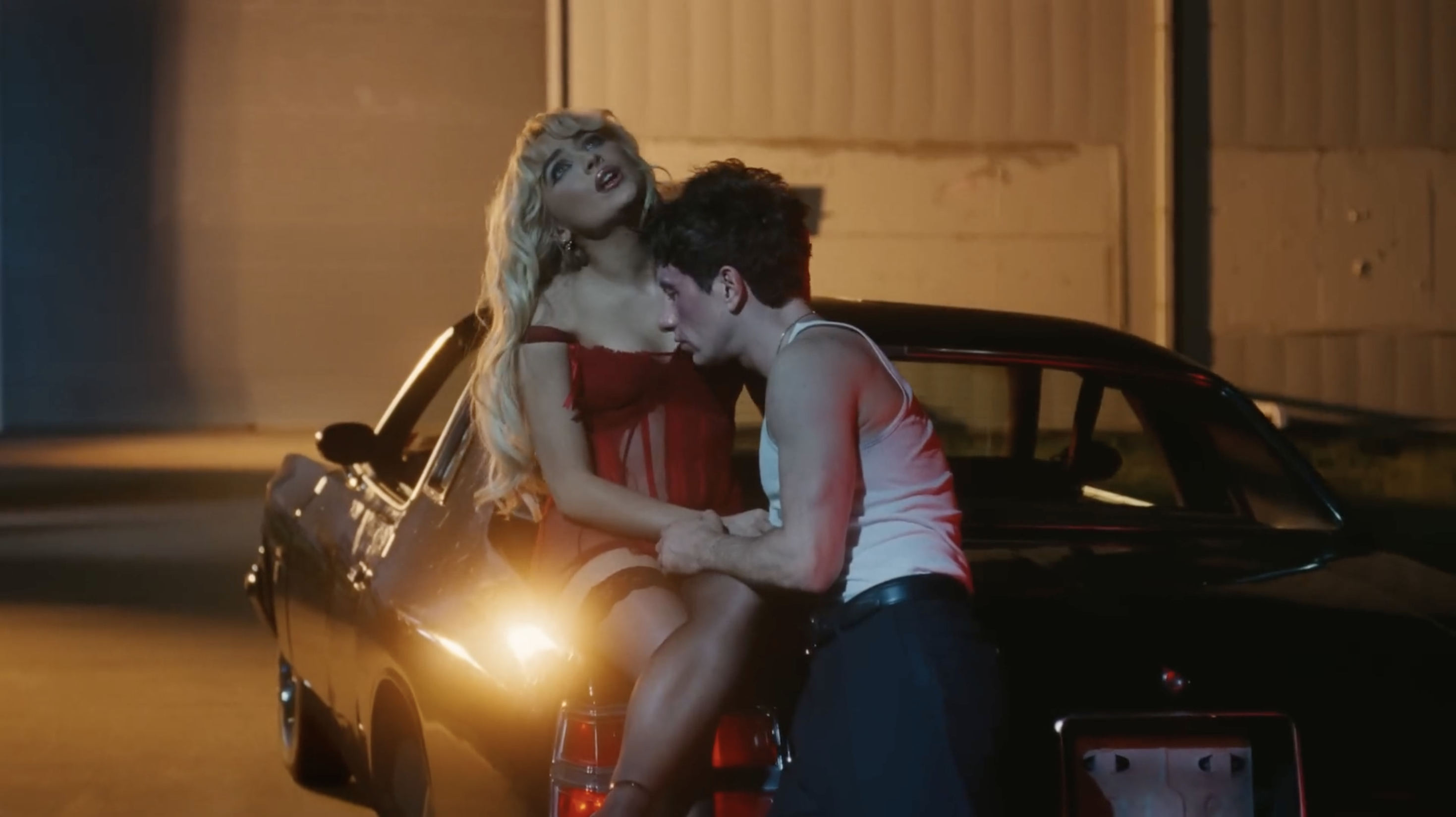 Sabrina sitting on a car while Barry leans in close to her in a scene from the music video