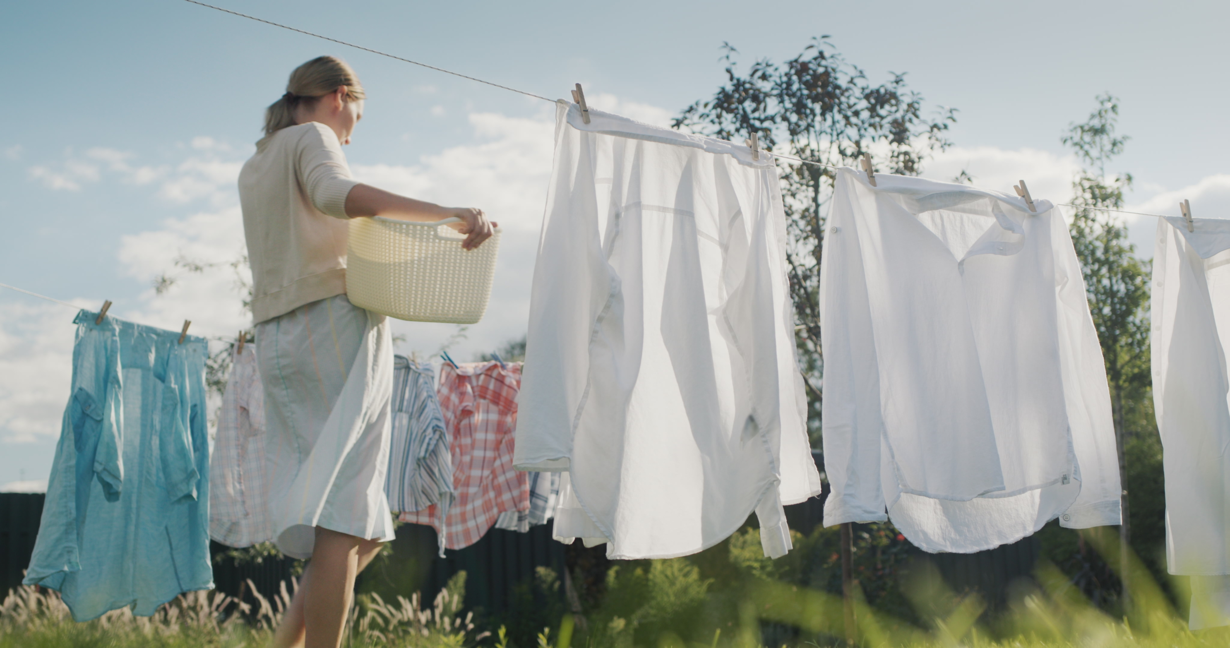 A person hangs freshly washed clothes on a clothesline in a sunny backyard, holding a laundry basket filled with clothes