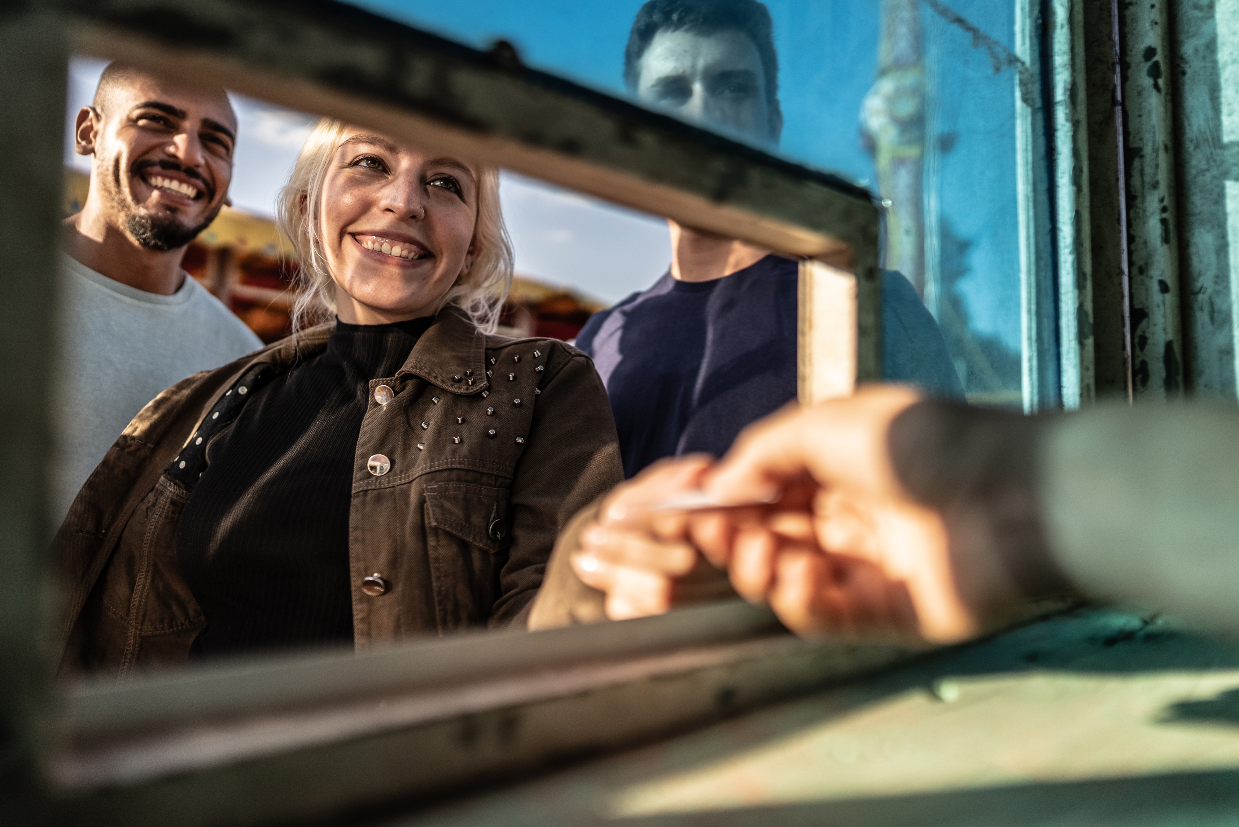 Three people are smiling as they approach a ticket booth window, with an arm extending a ticket to them