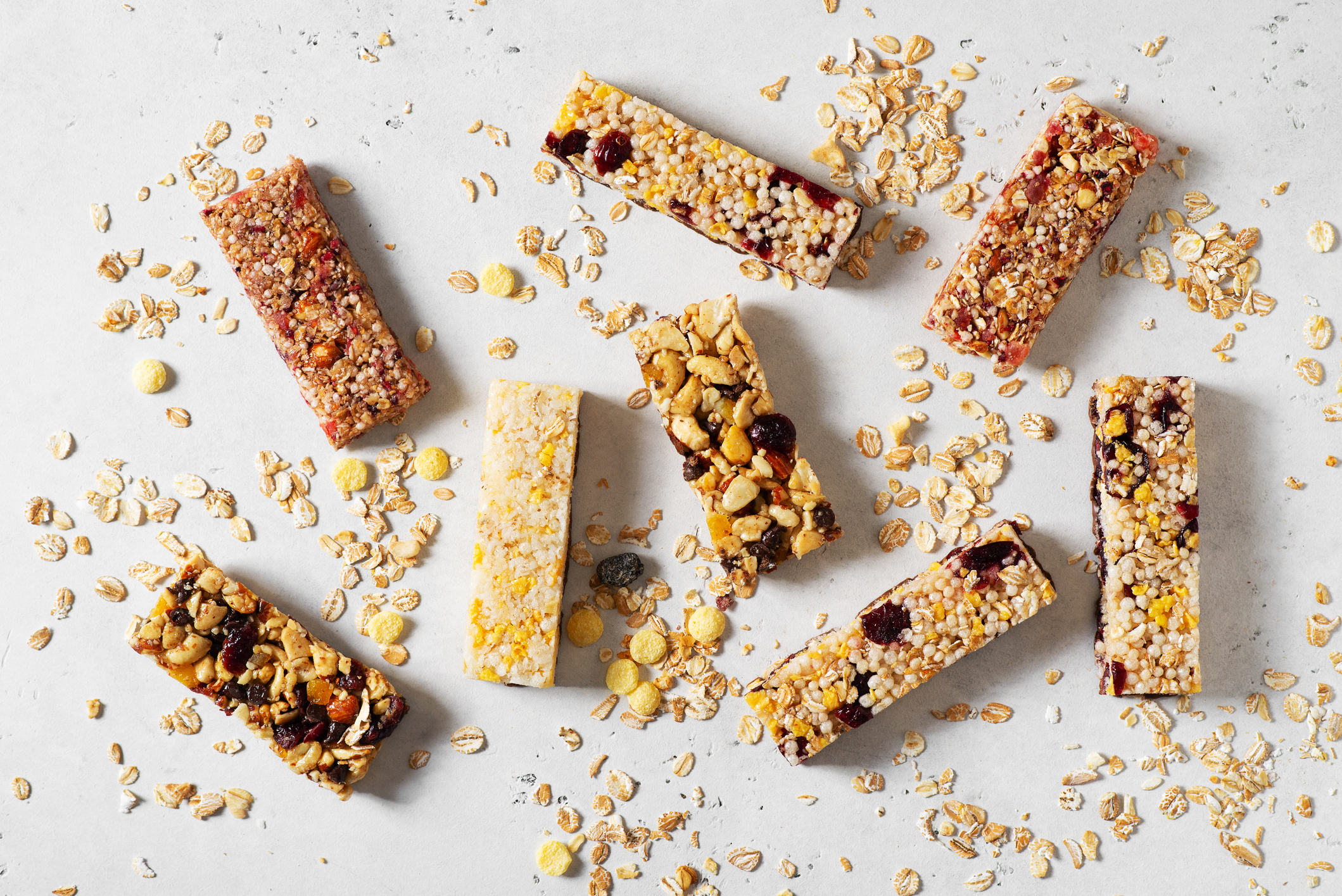 Various granola bars scattered among oats on a light surface. Bars contain nuts, dried fruit, and seeds