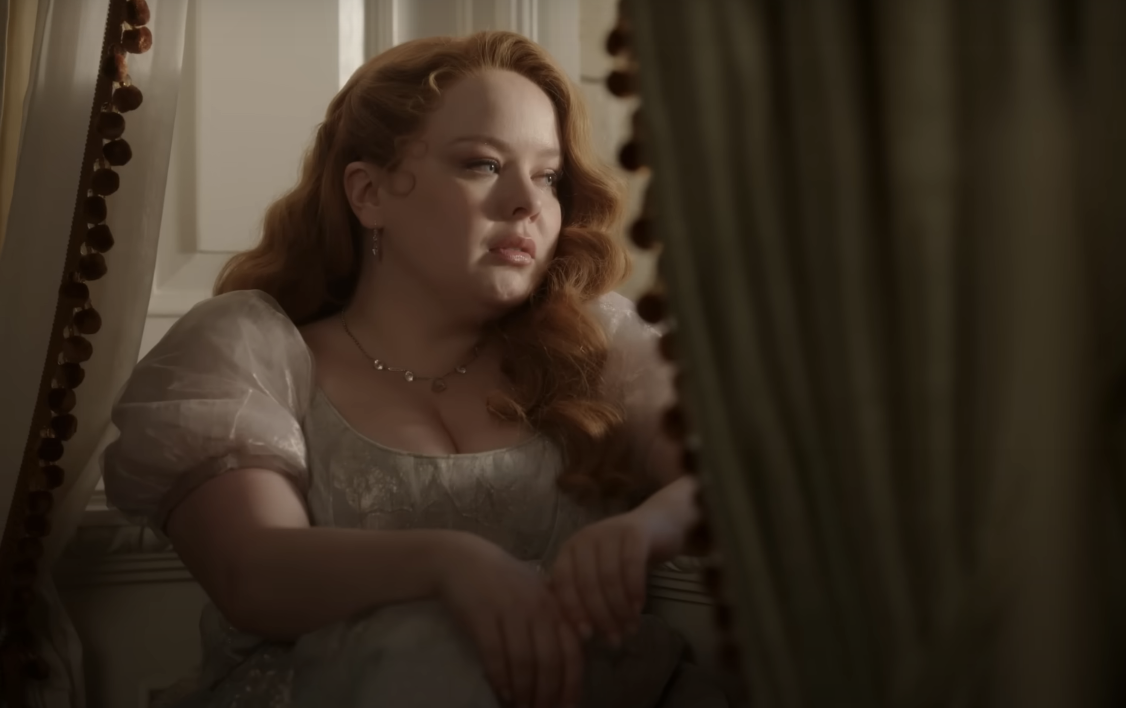 Nicola Coughlan sits by a window wearing a puff-sleeved period dress in a reflective and pensive pose with wavy hair cascading over her shoulders