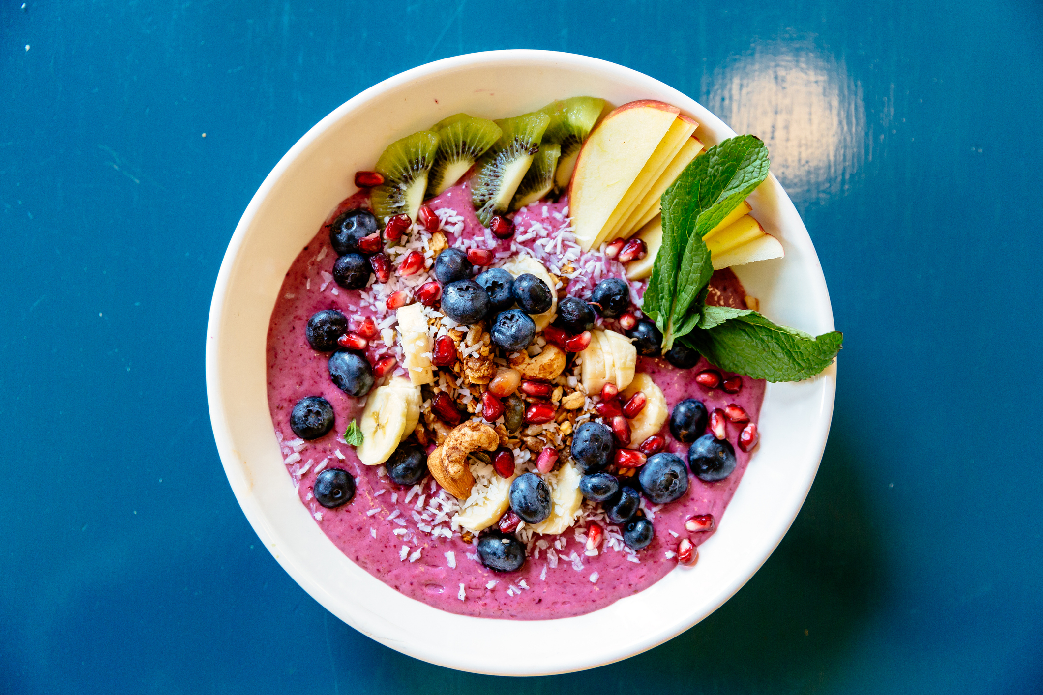 A bowl of smoothie topped with blueberries, sliced banana, apple slices, kiwi, pomegranate seeds, granola, shredded coconut, and mint leaves on a table