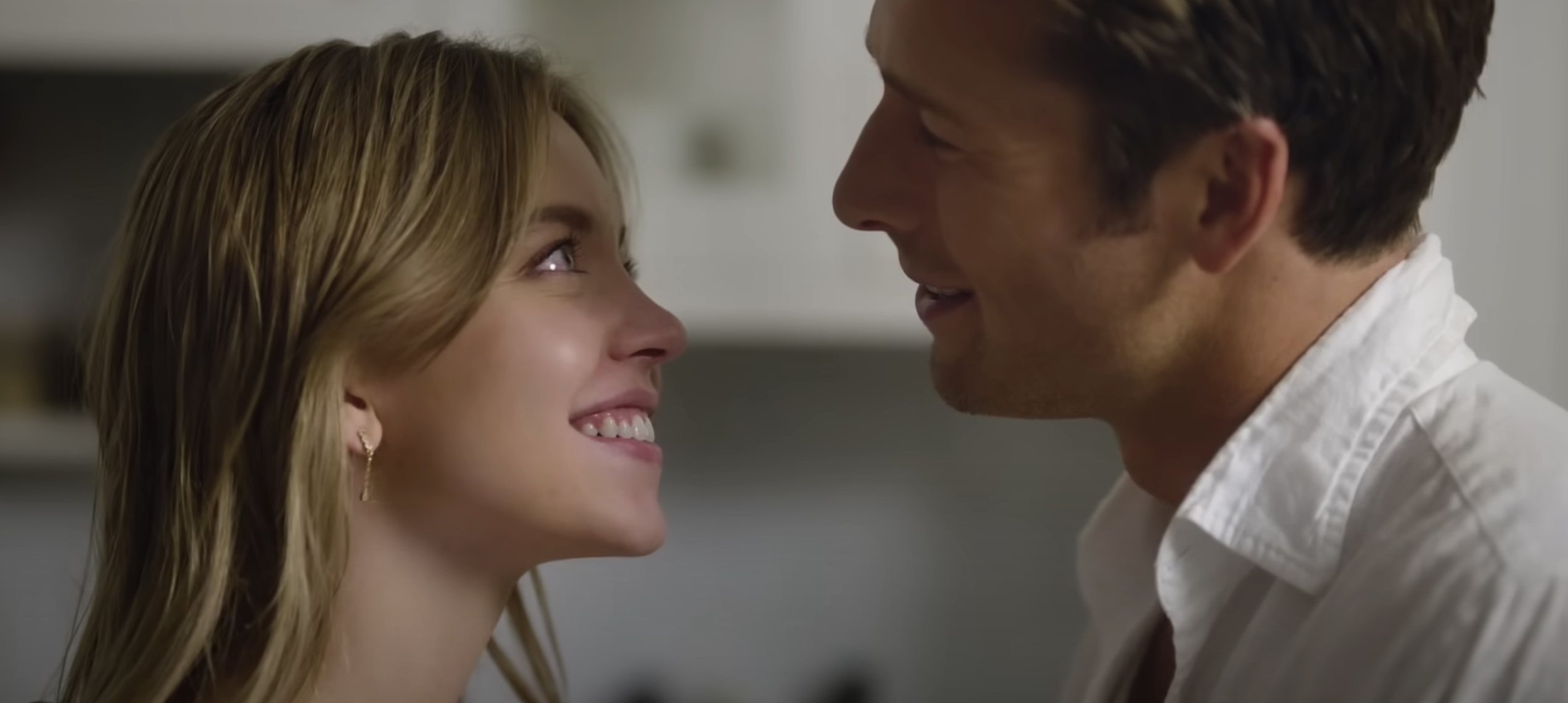 Sydney Sweeney and Glen Powell sharing a warm, smiling gaze at each other. Sydney wears a gold hoop earring, and Glen wears a white collared shirt