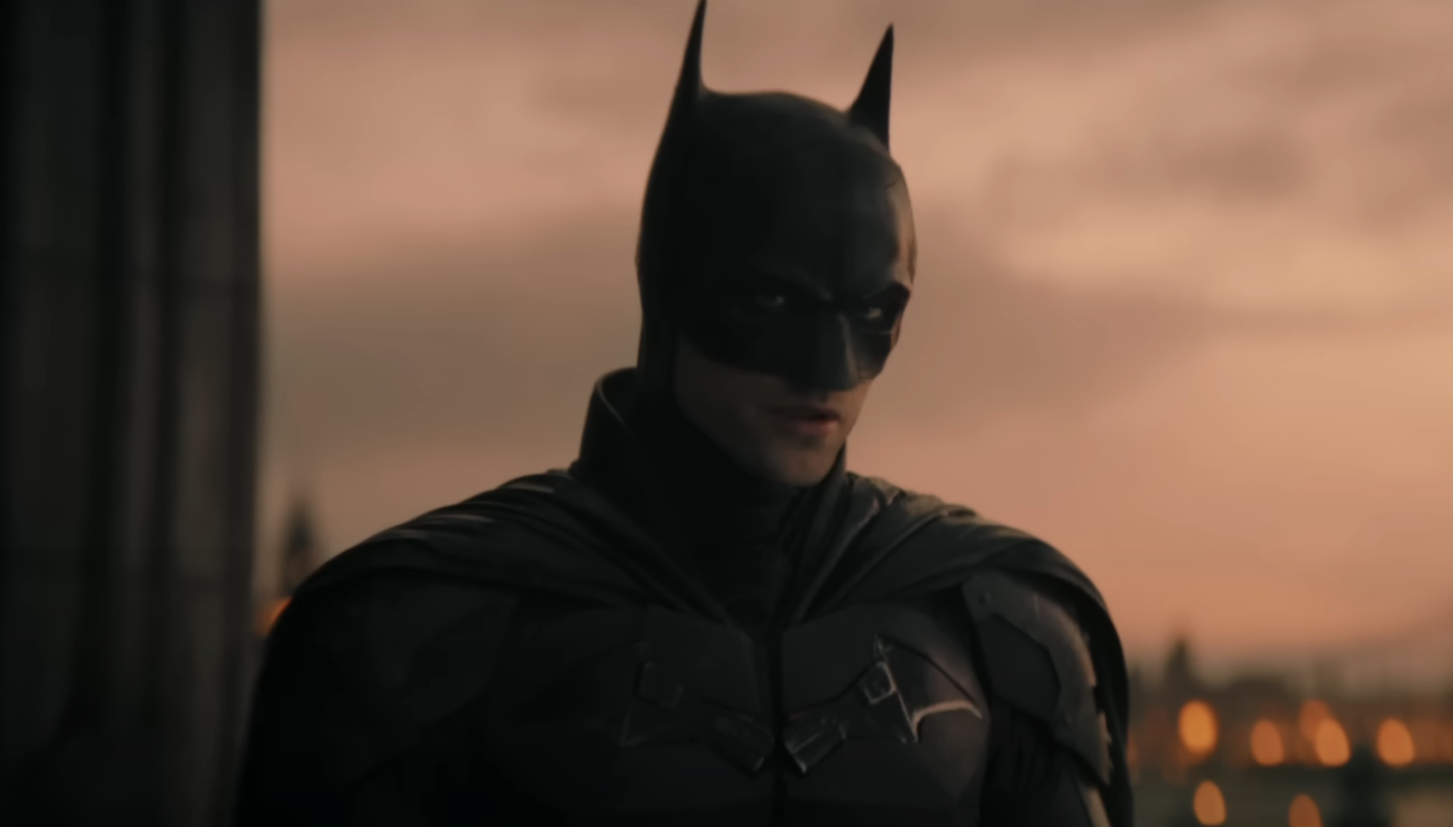 Batman in his suit stands against a dusk-lit cityscape