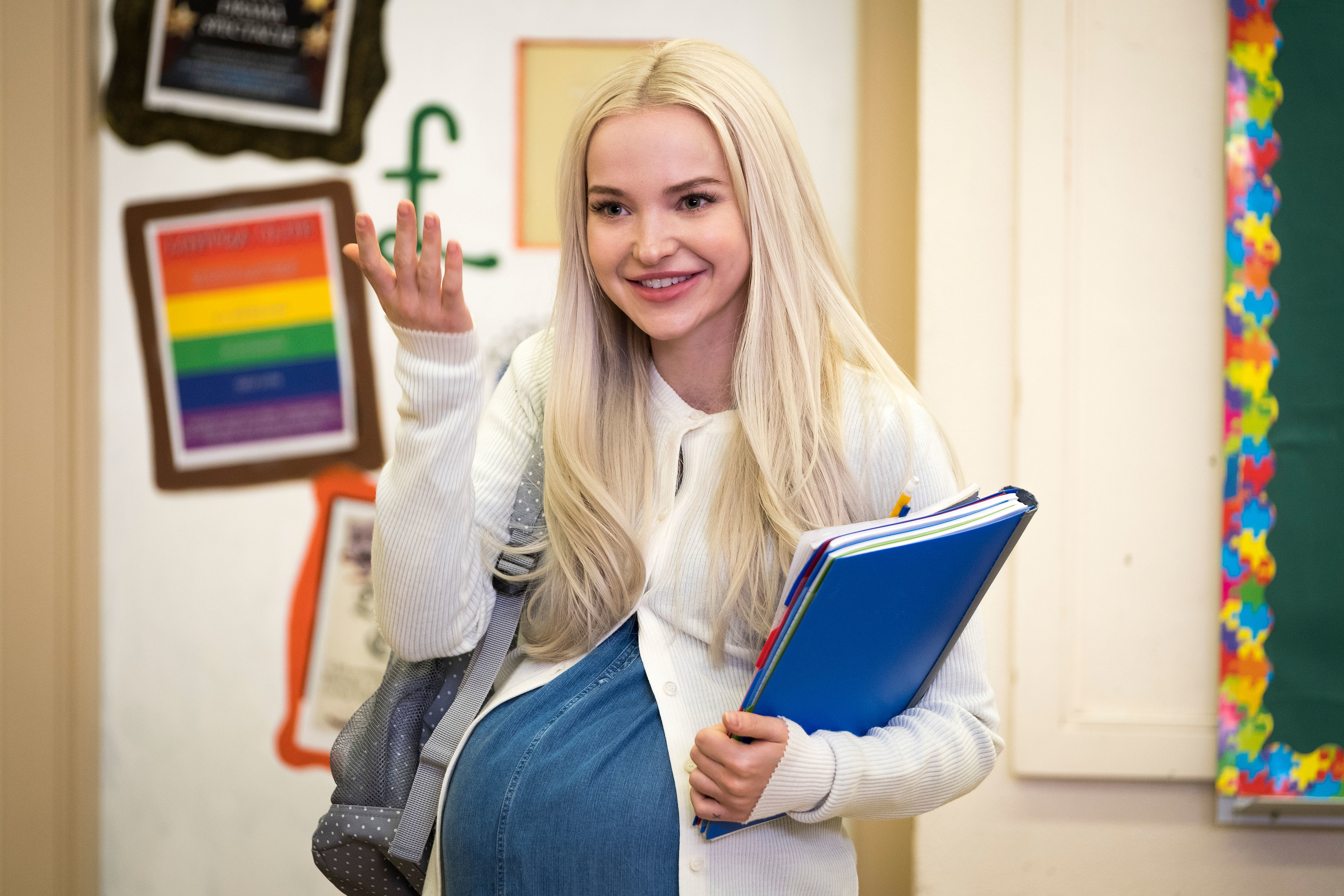 Dove Cameron is seen in a classroom setting, smiling and holding school books while wearing a white cardigan and a backpack