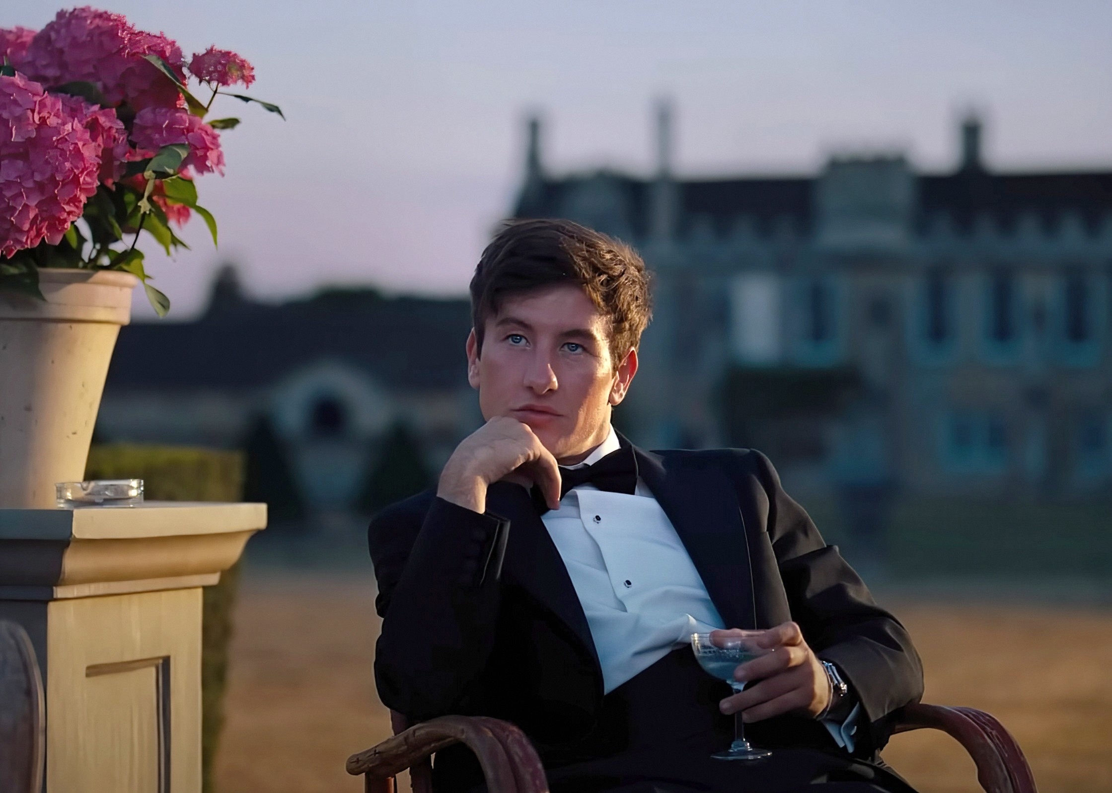 Barry Keoghan sits outdoors in a tuxedo, holding a glass, by a table with a flower pot. A grand building is seen in the background