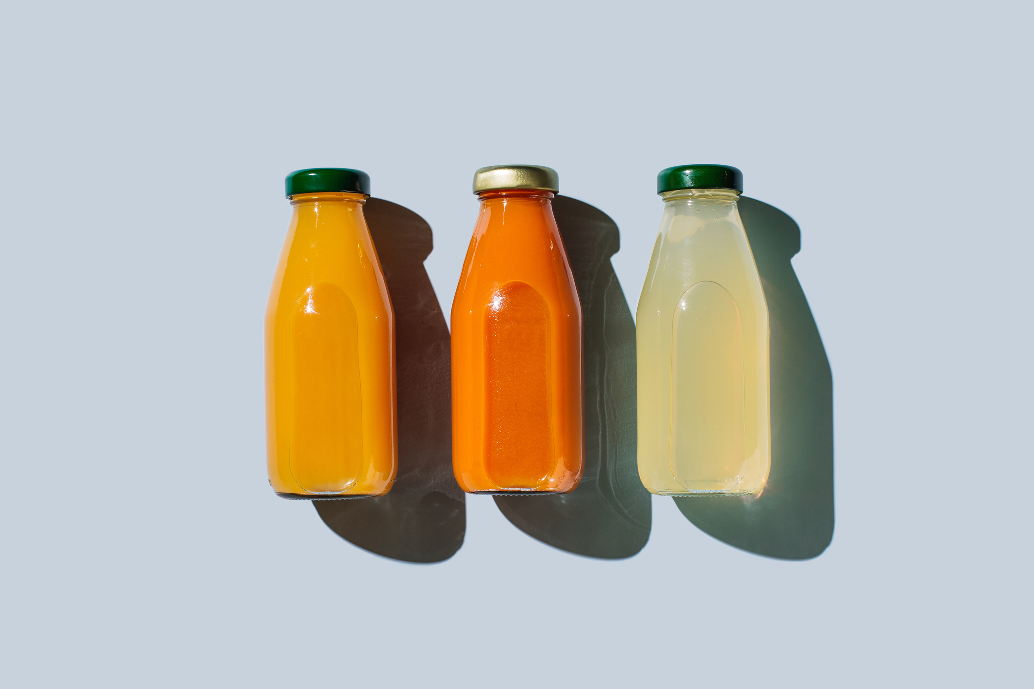 Three glass bottles with green caps contain orange, reddish-orange, and yellowish-clear liquids, placed in a row against a plain background