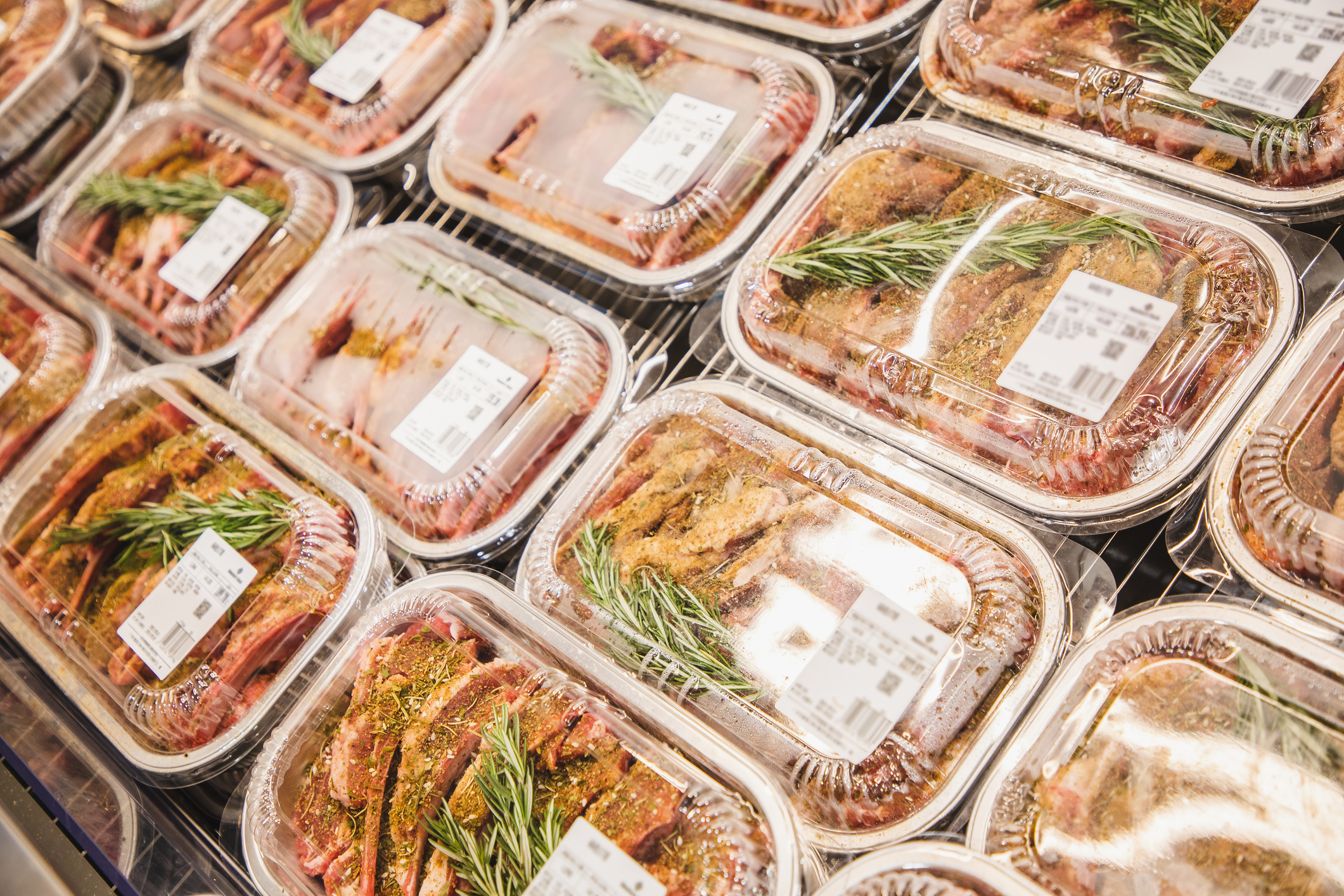 Packaged marinated meat with rosemary sprigs is displayed in a supermarket refrigerator section, each with a labeled price tag