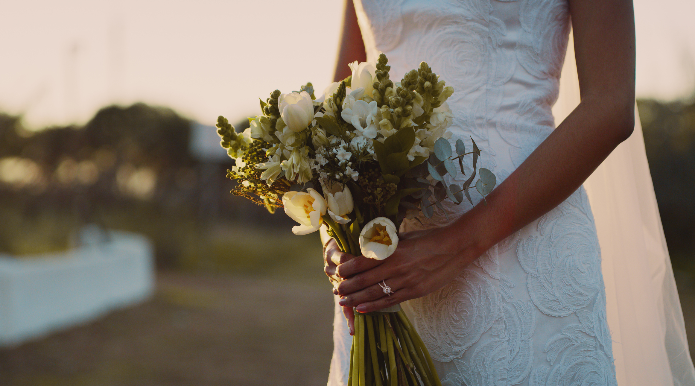 A person in a wedding dress holds a bouquet of flowers outdoors, with greenery in the background