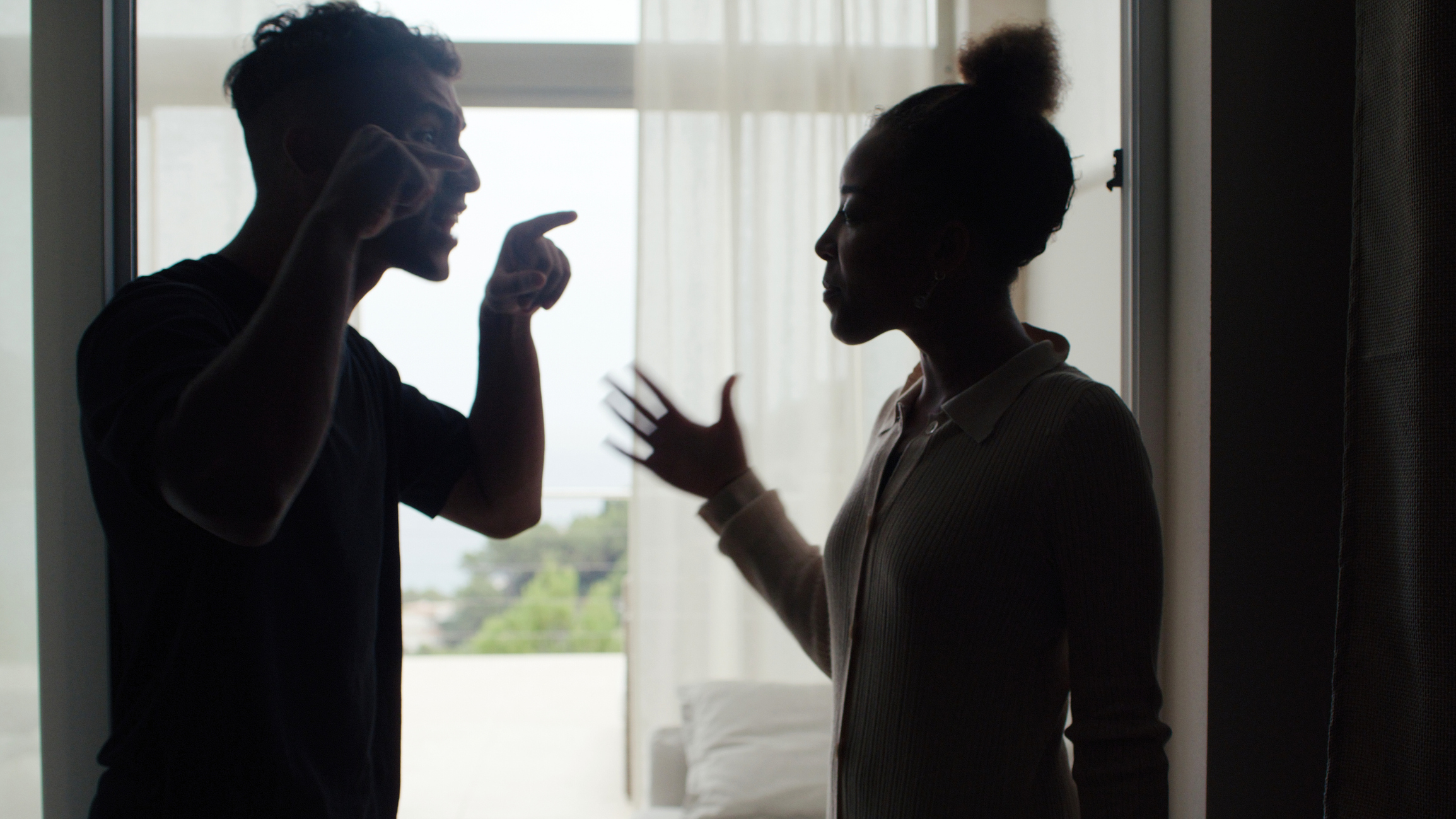 A man and woman are arguing indoors, with the man pointing at the woman. Silhouetted against a window, their emotions appear tense and confrontational