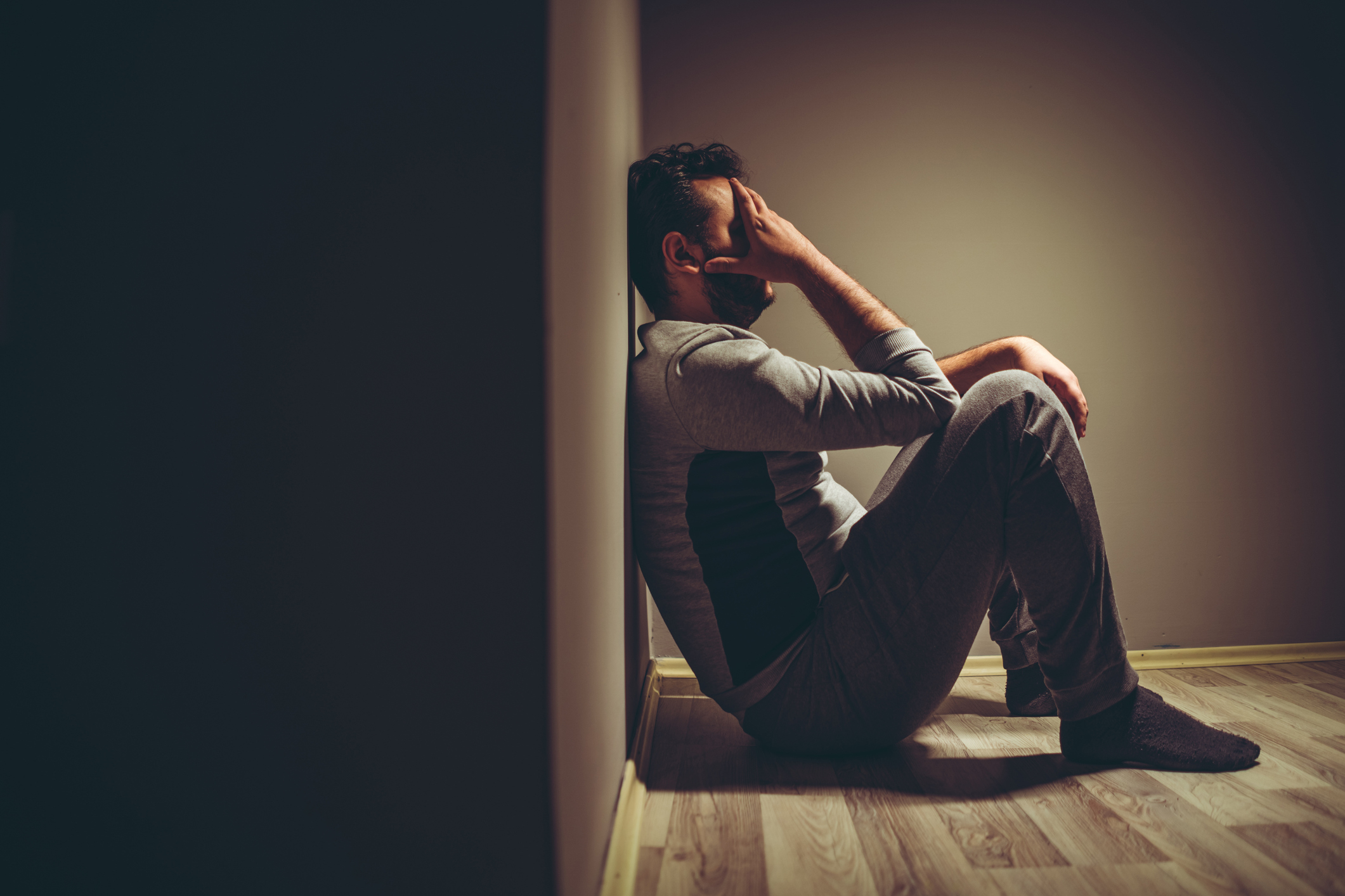 A person sits on the floor against a wall, head in hand, appearing distressed