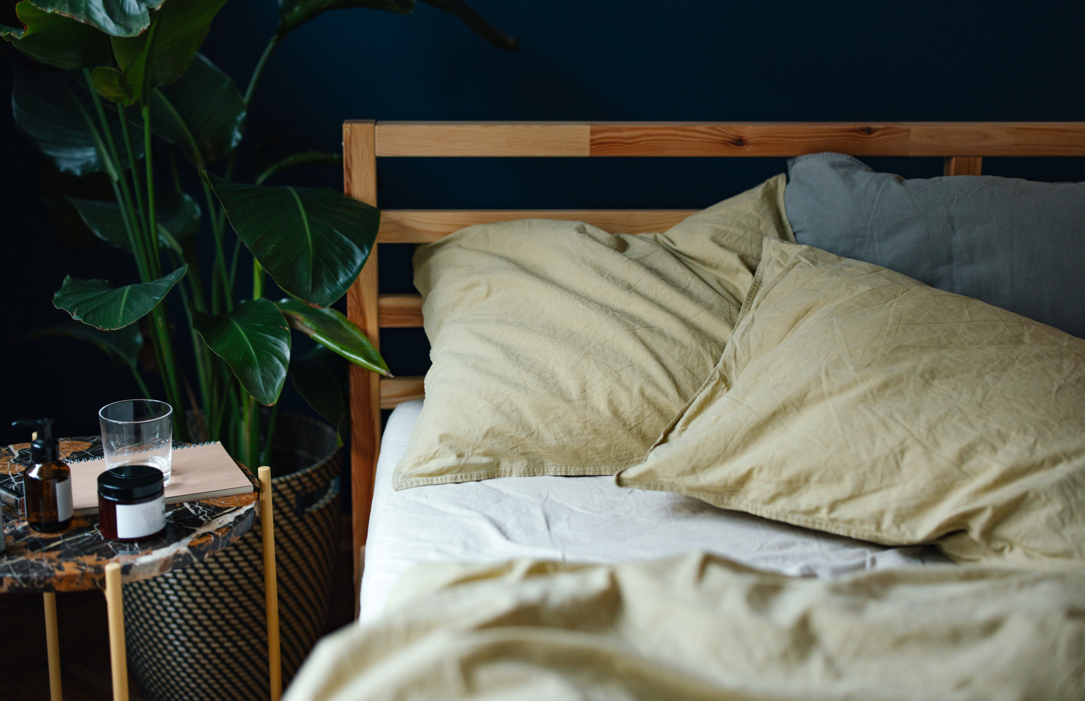 A cozy, unmade bed with beige sheets and pillows. There&#x27;s a side table with a glass of water, a plant, and some personal items