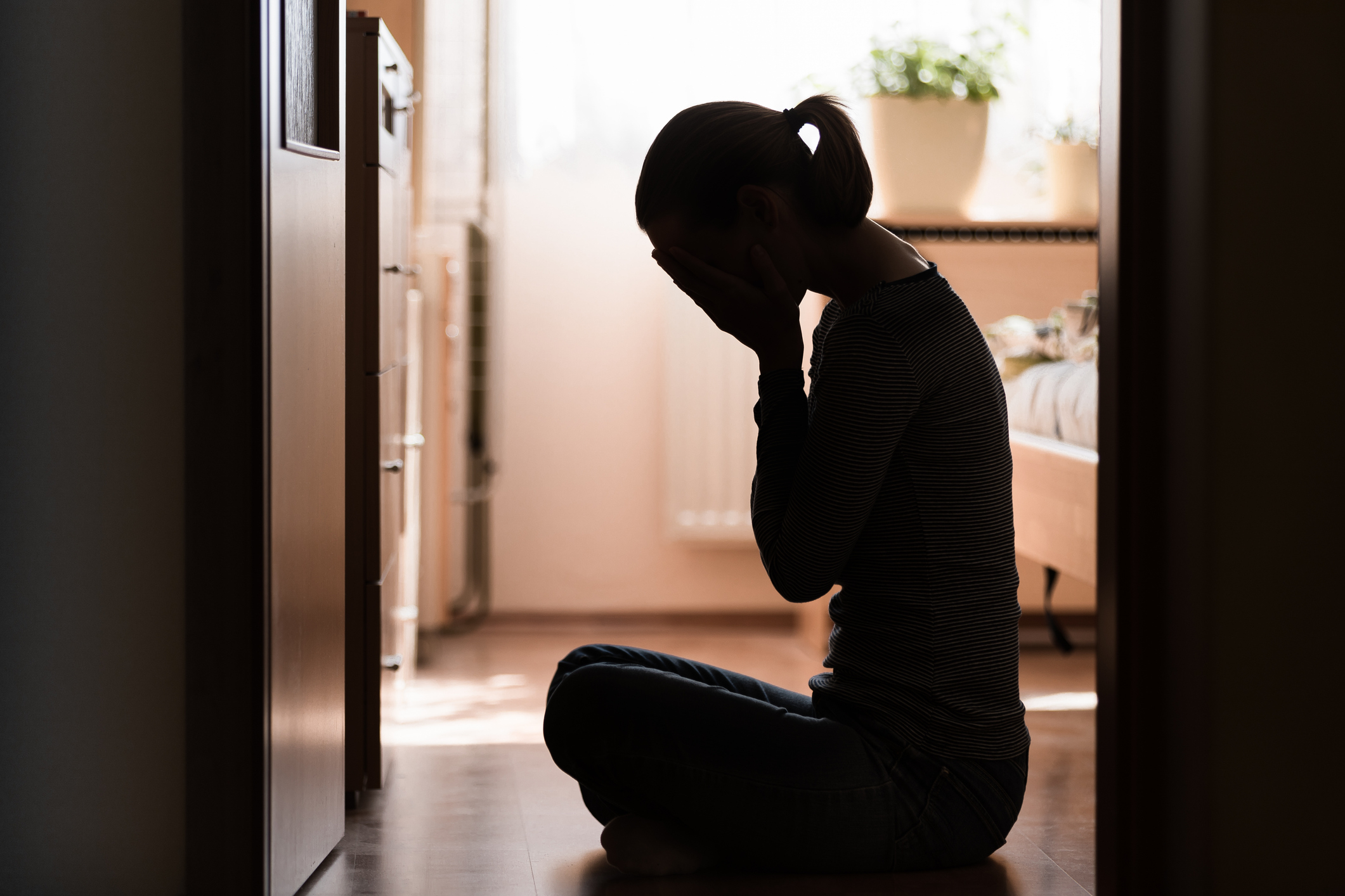 A person sits on the floor in a dimly lit room with their face in their hands, appearing distressed