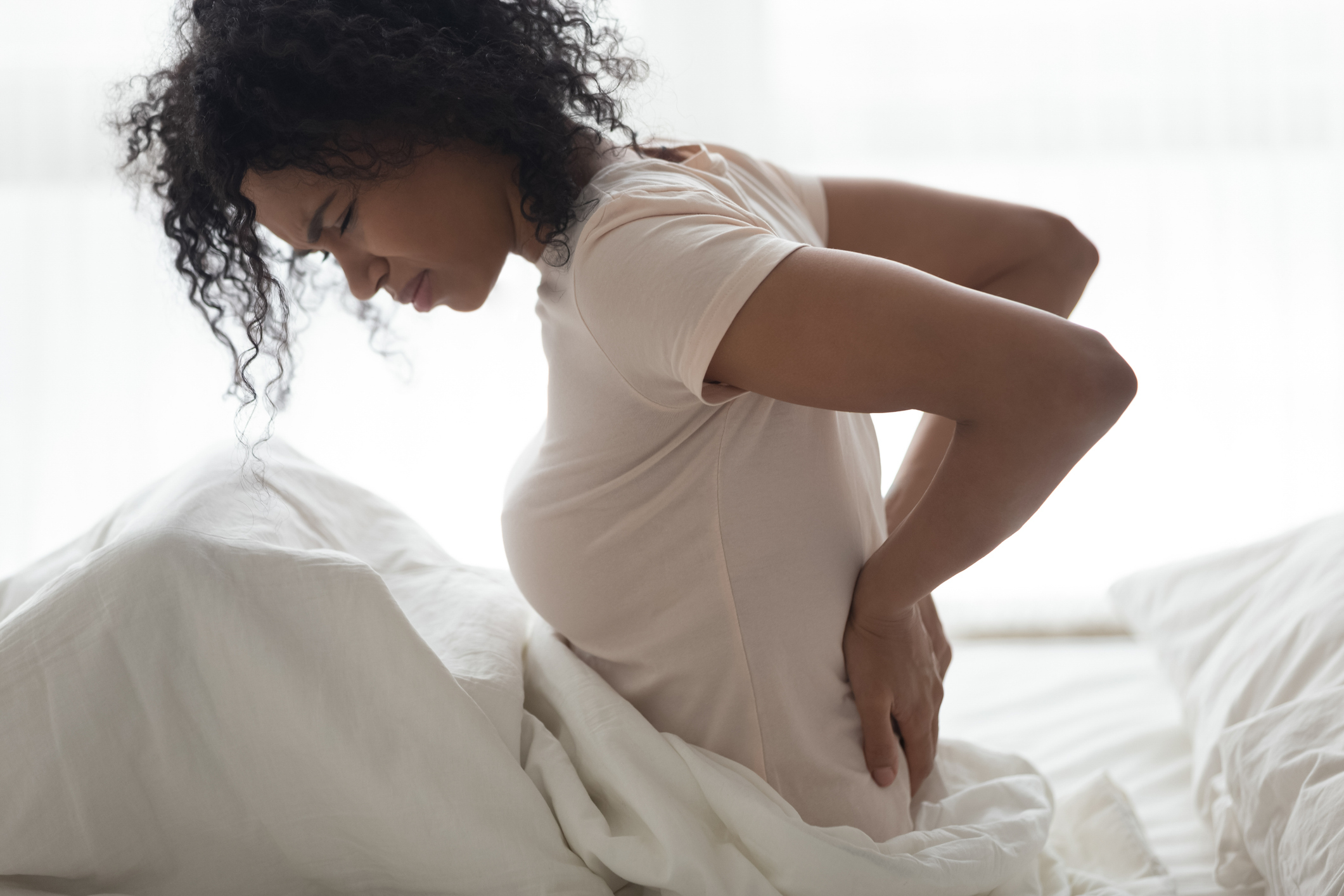 A person sitting on a bed, holding their lower back in pain with a slight grimace, suggesting discomfort or back pain