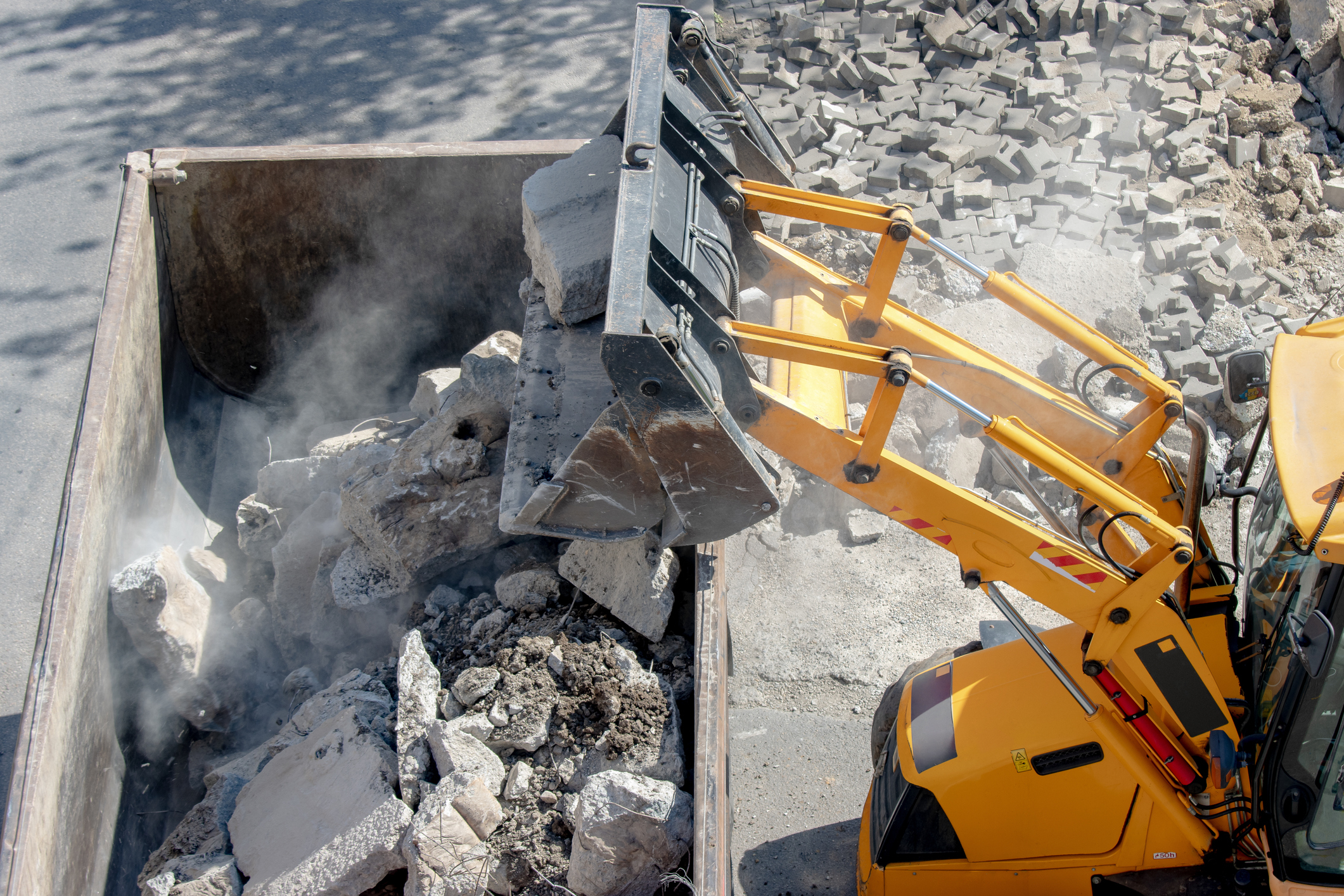 A yellow construction vehicle dumps a load of broken concrete and debris into a large metal container