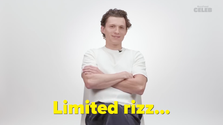 Tom Holland sits in a chair with arms folded, wearing a casual light shirt and dark pants. The text at the bottom reads, &quot;Limited rizz...&quot;