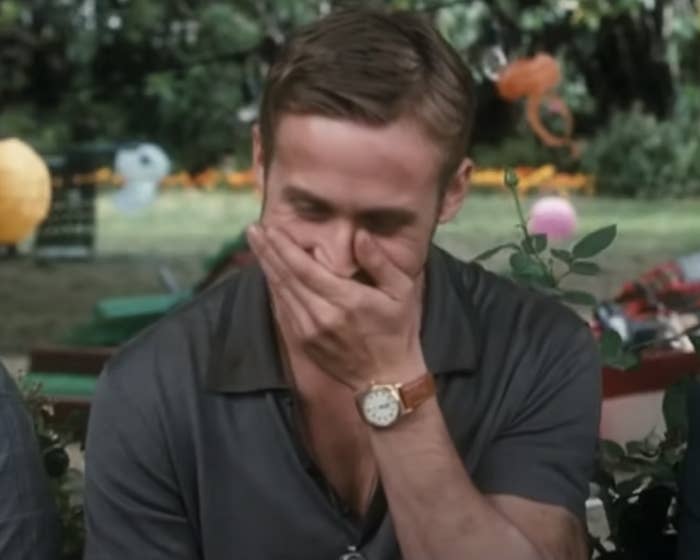 Ryan Gosling covers his mouth while laughing in an outdoor setting decorated with festive ornaments