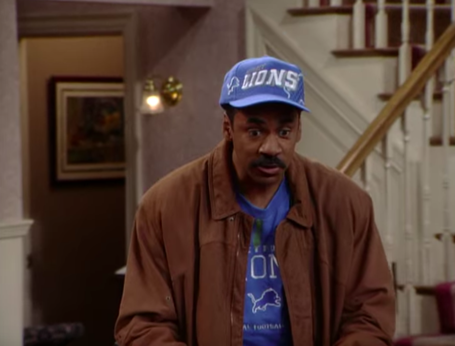 Man in a casual outfit with a blue Detroit Lions cap stands inside a house near a staircase