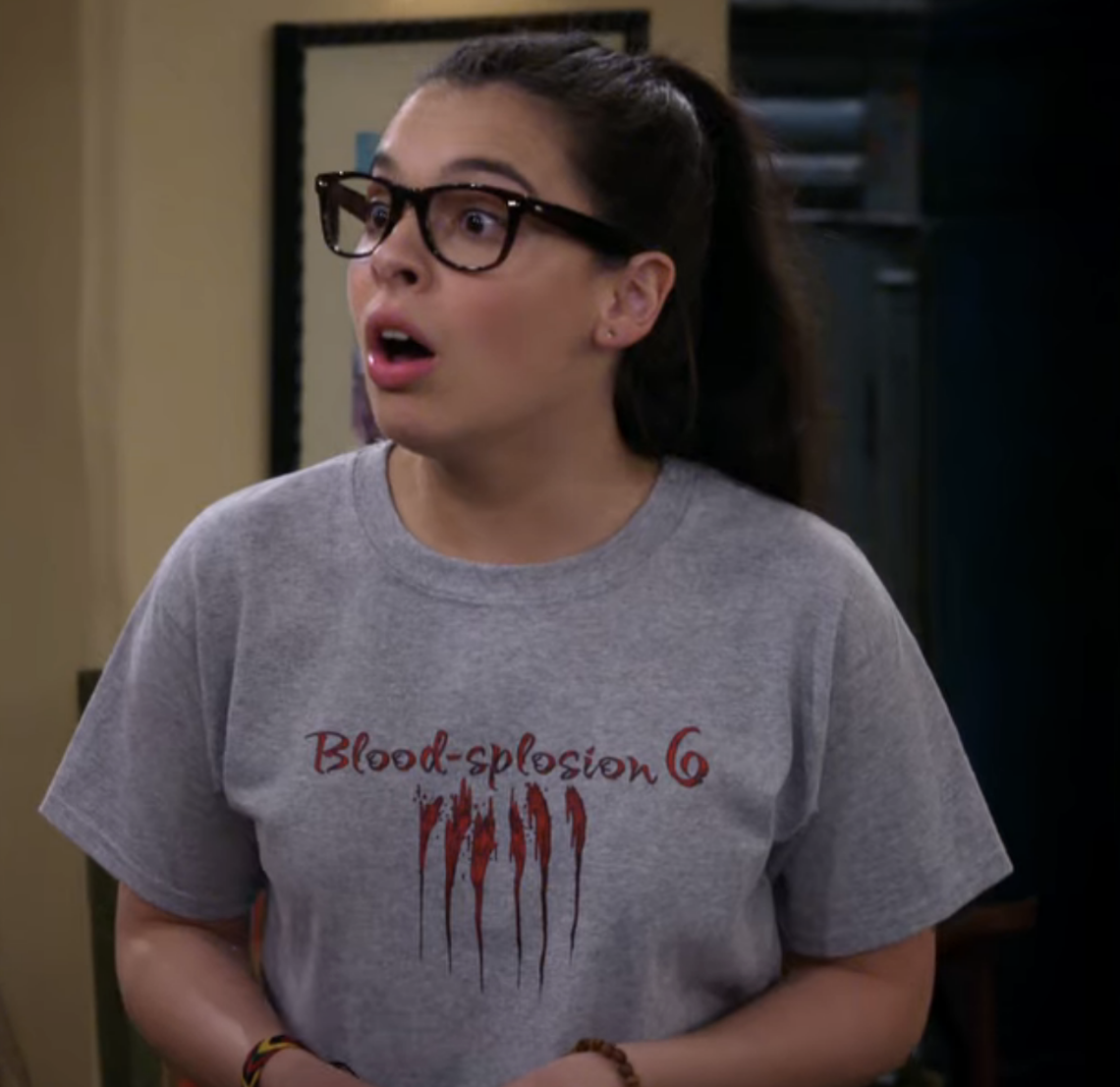 Young woman with long hair in glasses, wearing a grey &quot;Blood-splosion 6&quot; t-shirt, looks surprised