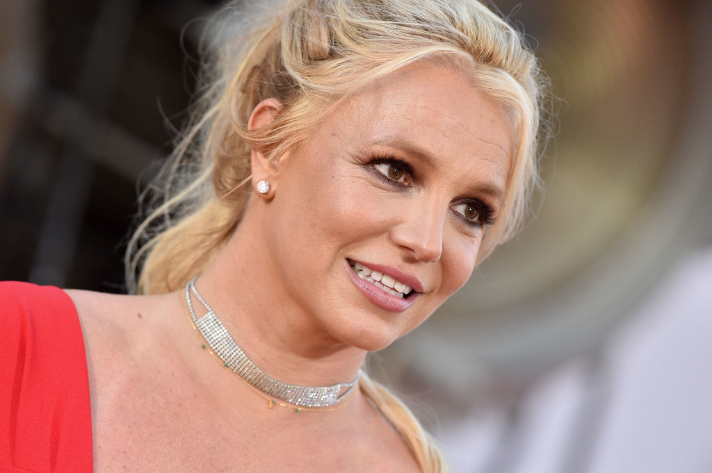 Britney Spears on a red carpet, wearing an elegant sleeveless dress and a layered choker necklace, with her hair styled in a loose updo