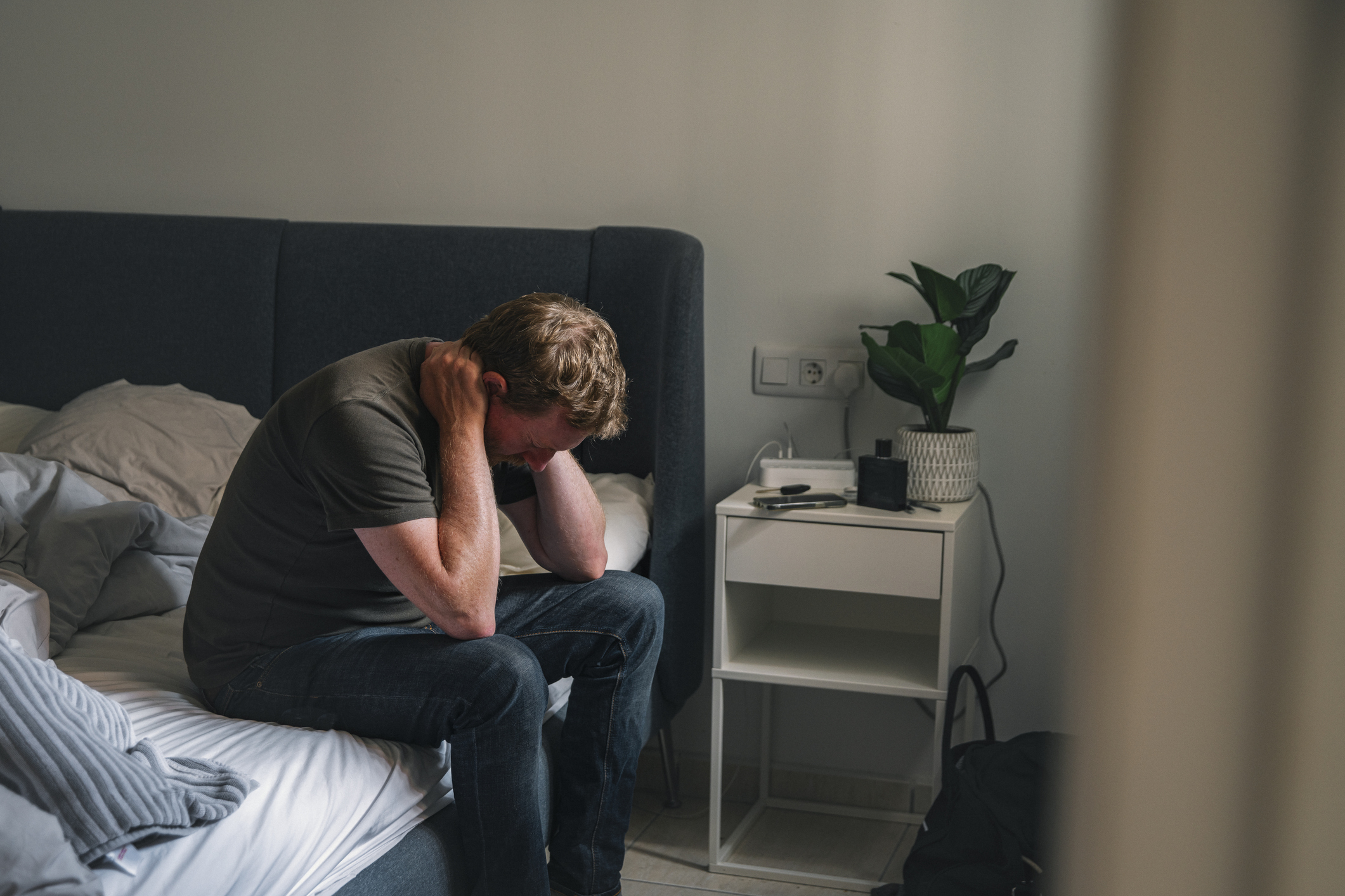 A person sits on a bed with their head in their hands, appearing distressed, in a bedroom setting with a nightstand, plant, and personal items