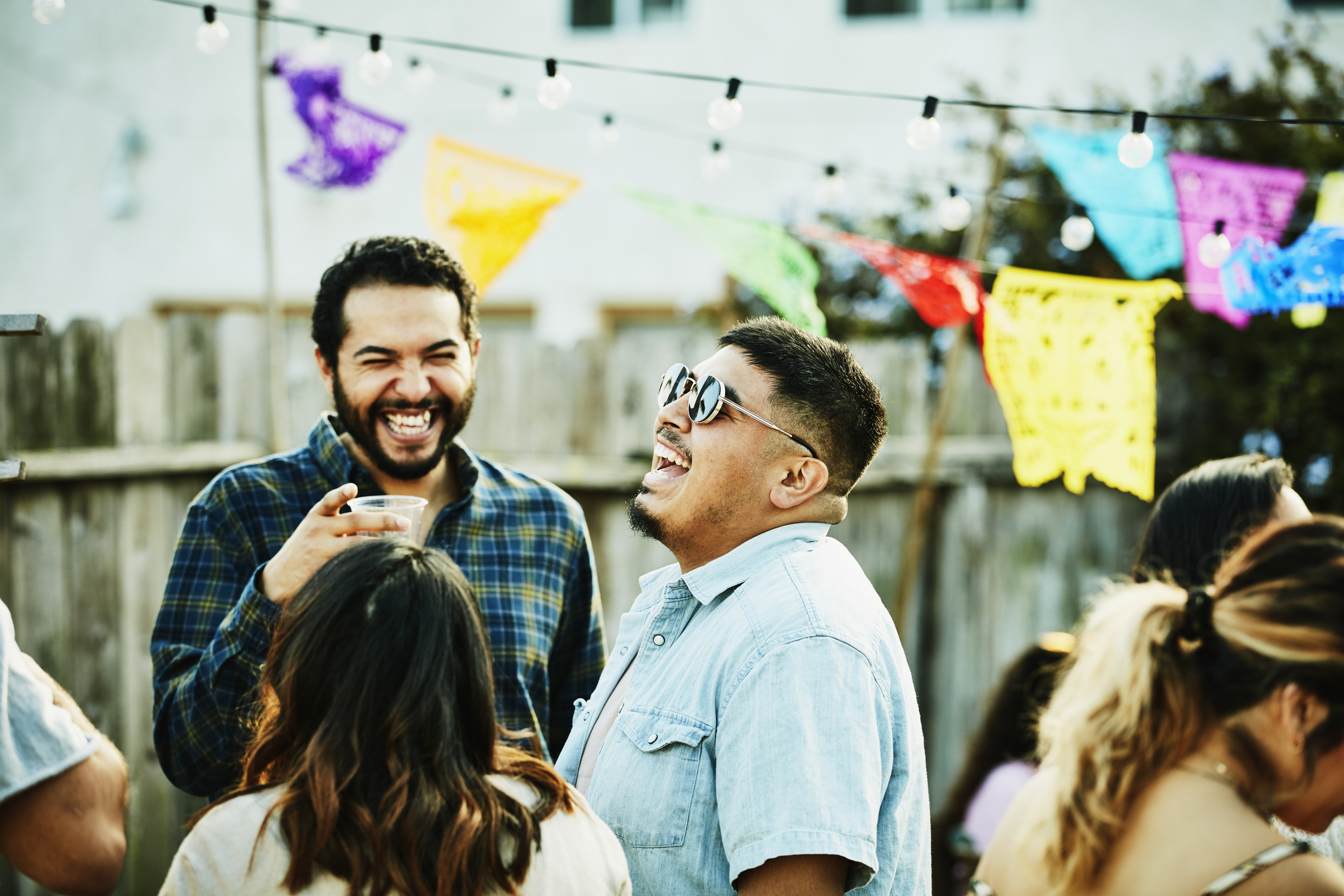 A group of people are laughing and mingling at an outdoor gathering with colorful festive banners in the background