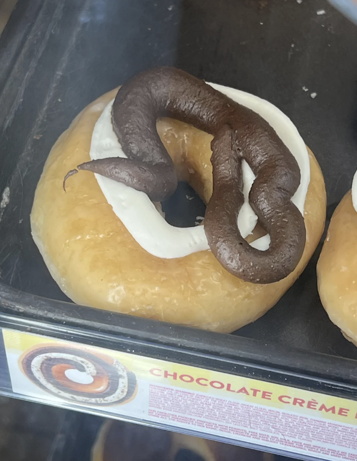 A doughnut with white icing is topped with what appears to be chocolate frosting, shaped like feces. The label reads &quot;CHOCOLATE CRÈME.&quot;