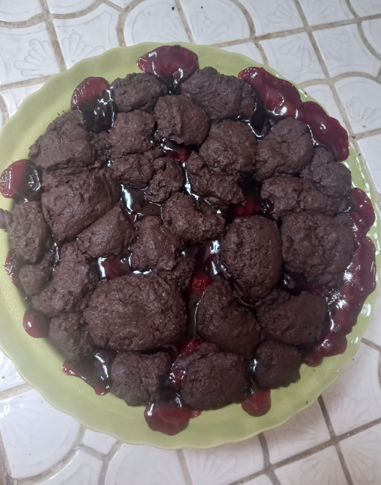 A cherry cobbler with dark, chocolate-like dumplings on top on a green plate placed on a tiled surface