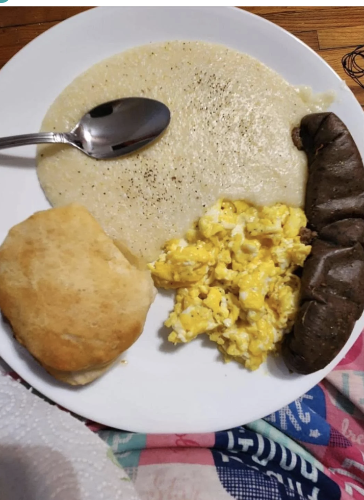 A plate of food with grits, scrambled eggs, a biscuit, and a sausage link. A spoon is placed within the grits