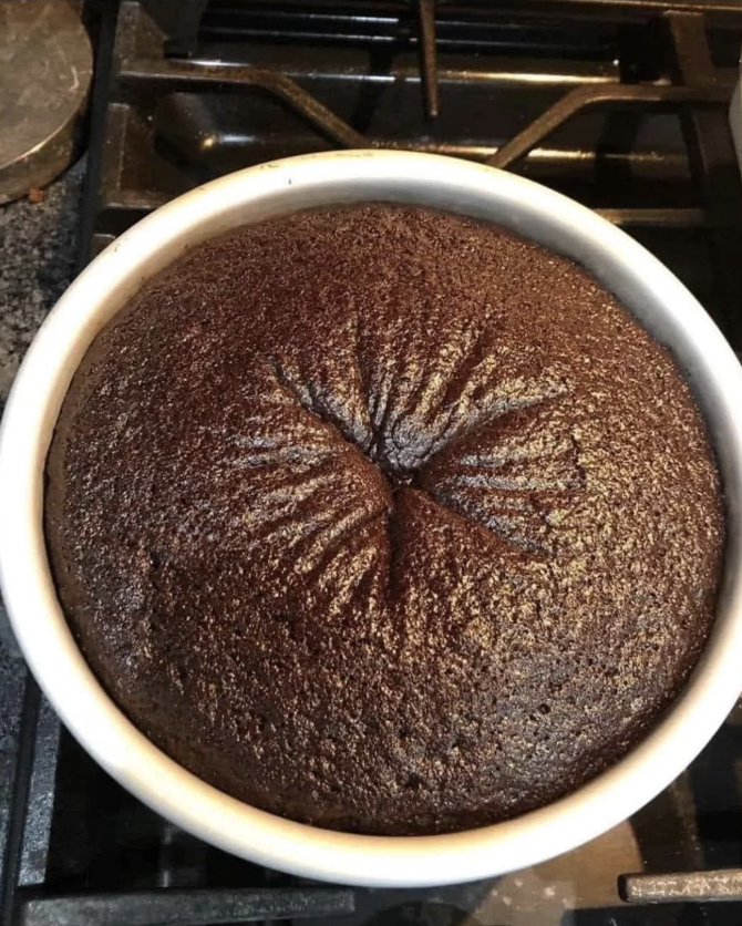A round chocolate cake freshly baked in a circular baking tin on a stovetop. The cake has a textured surface and a central indentation
