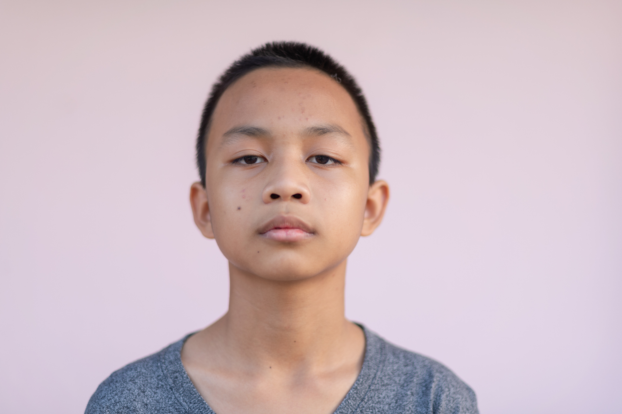 A young person with short hair and a neutral expression wearing a simple grey shirt stands against a plain background