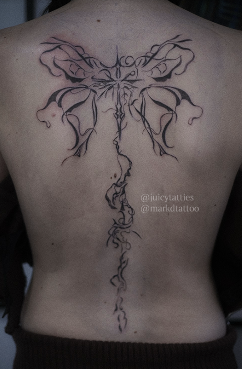 A photo of a person&#x27;s back with a detailed tattoo of intricate, symmetrical designs resembling wings and flowing lines down the spine. Text reads &quot;@juicytatties @markdtattoo&quot;