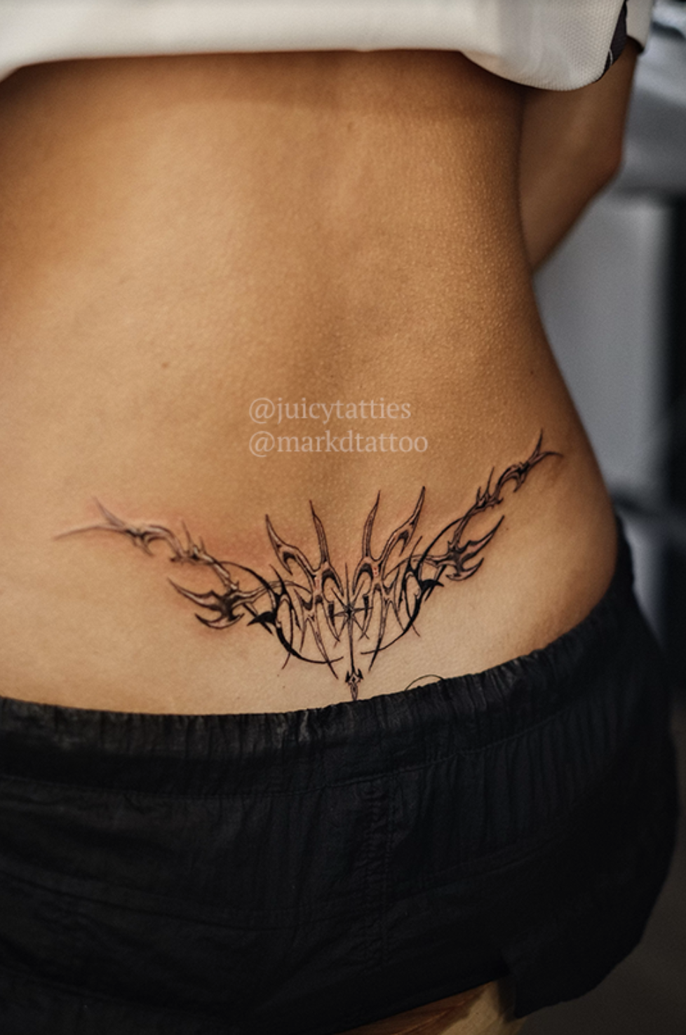 Lower back tattoo featuring an artistic abstract design with the text &quot;@juicytatties @markdtattoo&quot; written above the tattoo