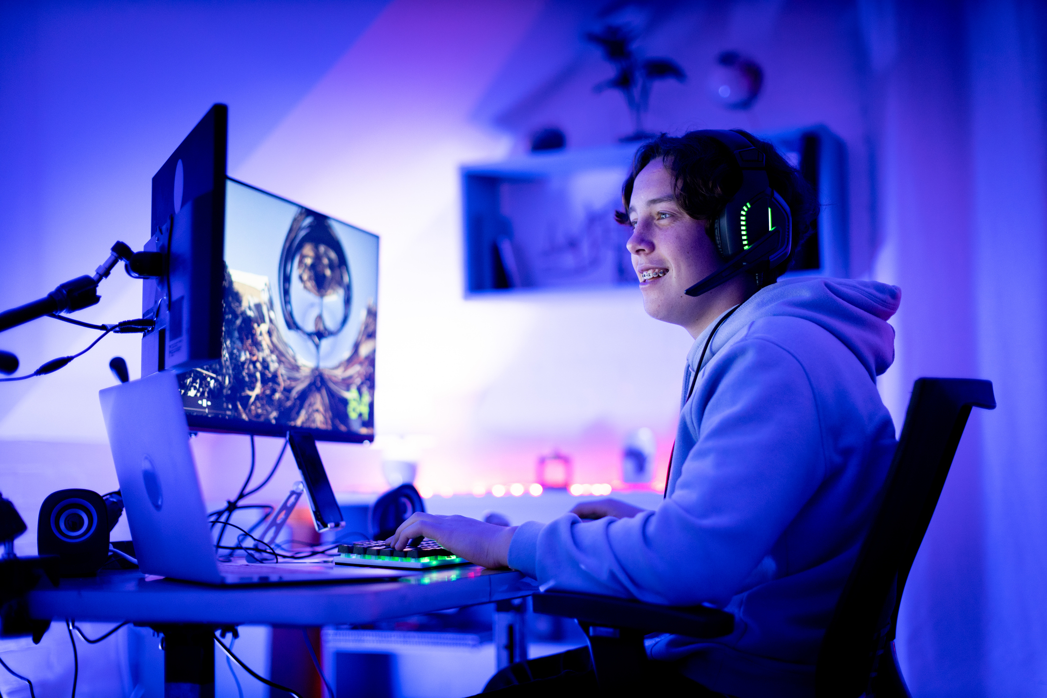 A person wearing a hoodie and a gaming headset sits at a desk with multiple monitors, actively engaging in a video game. The room background includes shelves and tech equipment