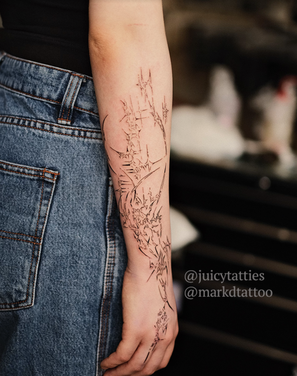 A person’s arm is shown with a detailed black line tattoo, mostly dragon-themed, just above the wrist. The tattoo artist&#x27;s social media tags, @juicytatties and @markdtattoo, are visible