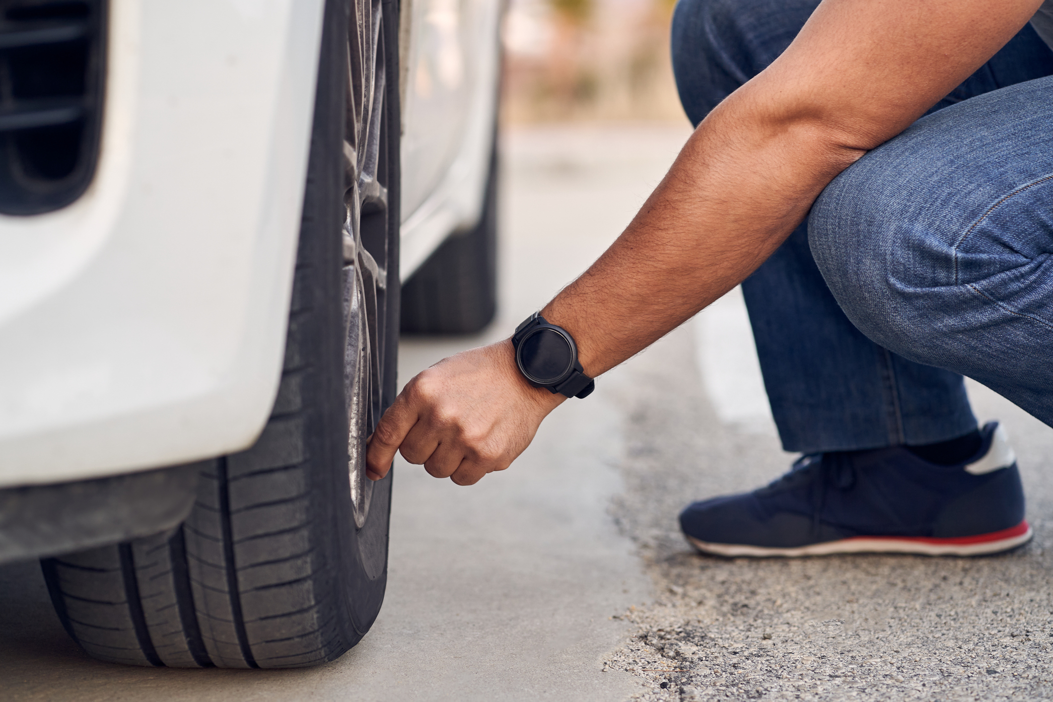 A person in casual clothing examines the tread on a tire of a parked car