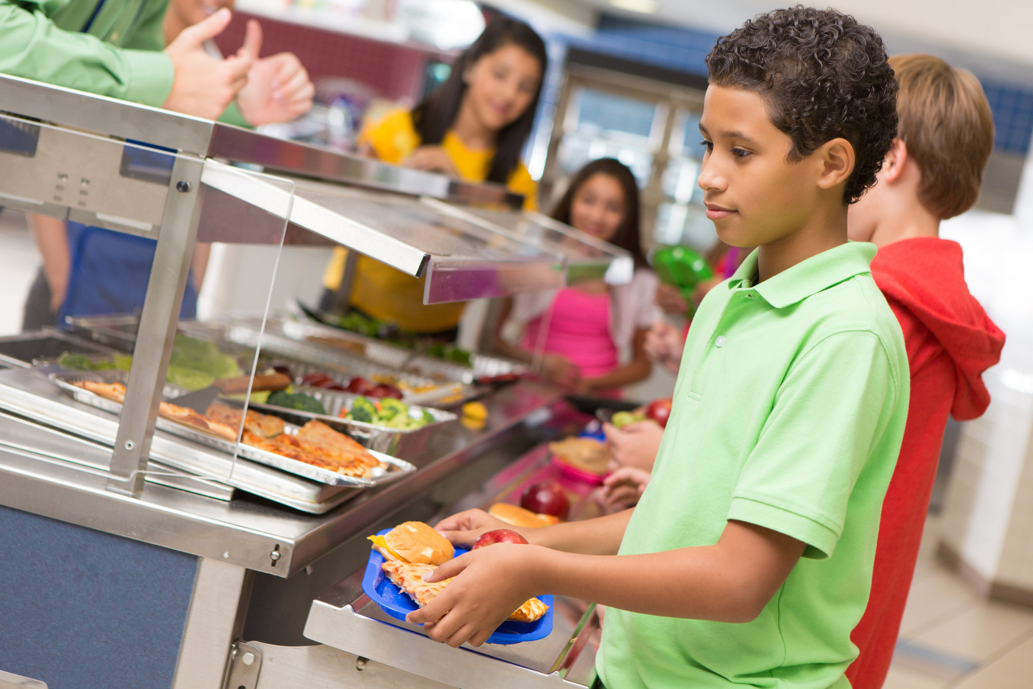 Students waiting in line at a school cafeteria, selecting food from various trays with assistance from staff
