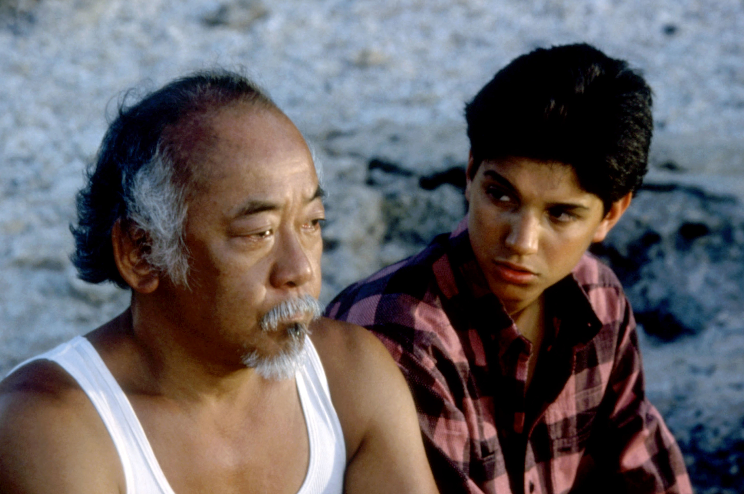 Pat Morita and Ralph Macchio in a scene from &quot;The Karate Kid,&quot; with Morita in a white tank top and Macchio in a plaid shirt, both looking serious