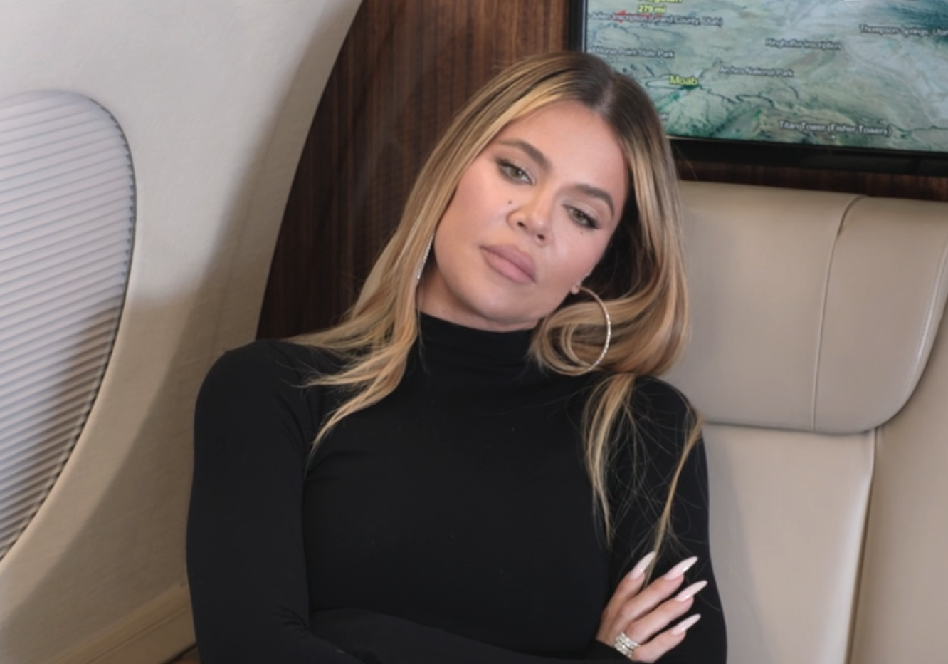 Khloé Kardashian is sitting in an airplane seat, wearing a black turtleneck long-sleeve shirt with her arms crossed, looking thoughtful