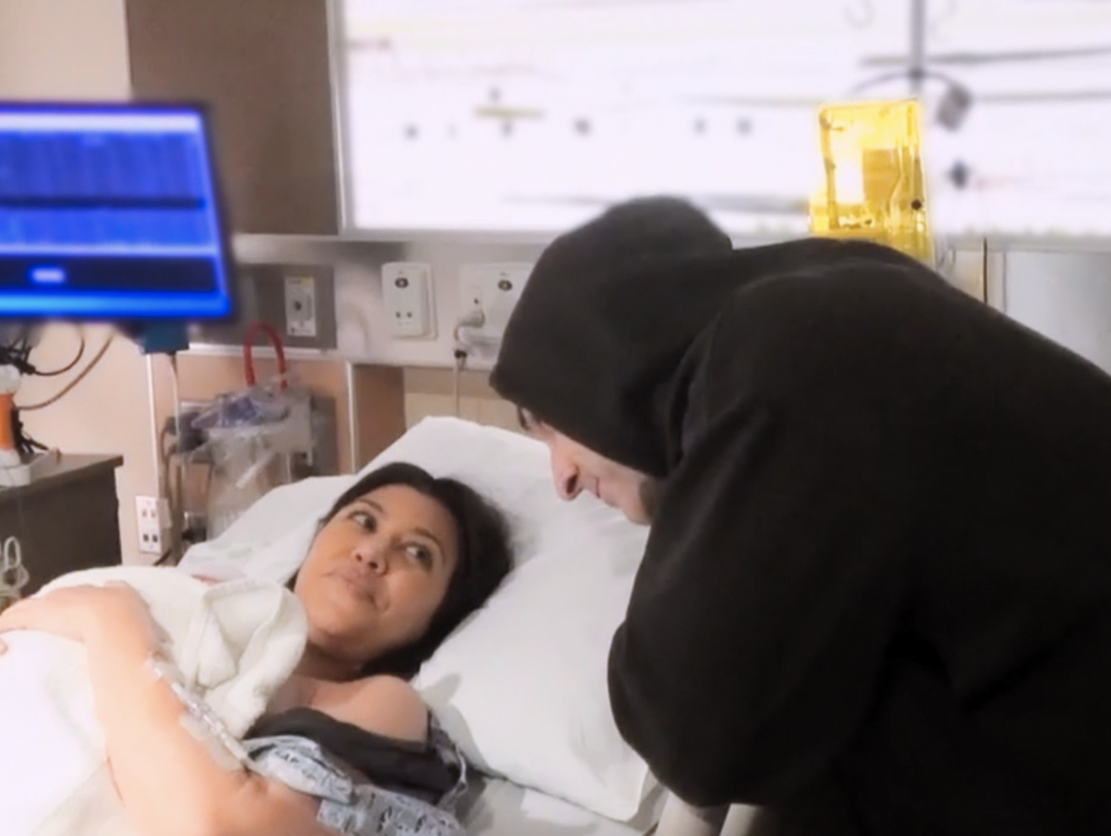 Kourtney Kardashian lies in a hospital bed holding a baby as Travis Barker, wearing a hooded sweater, leans close to her