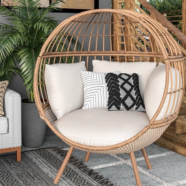 Wicker egg chair with cream cushions and two patterned throw pillows, placed in a cozy outdoor sitting area with plants and wooden furnishings